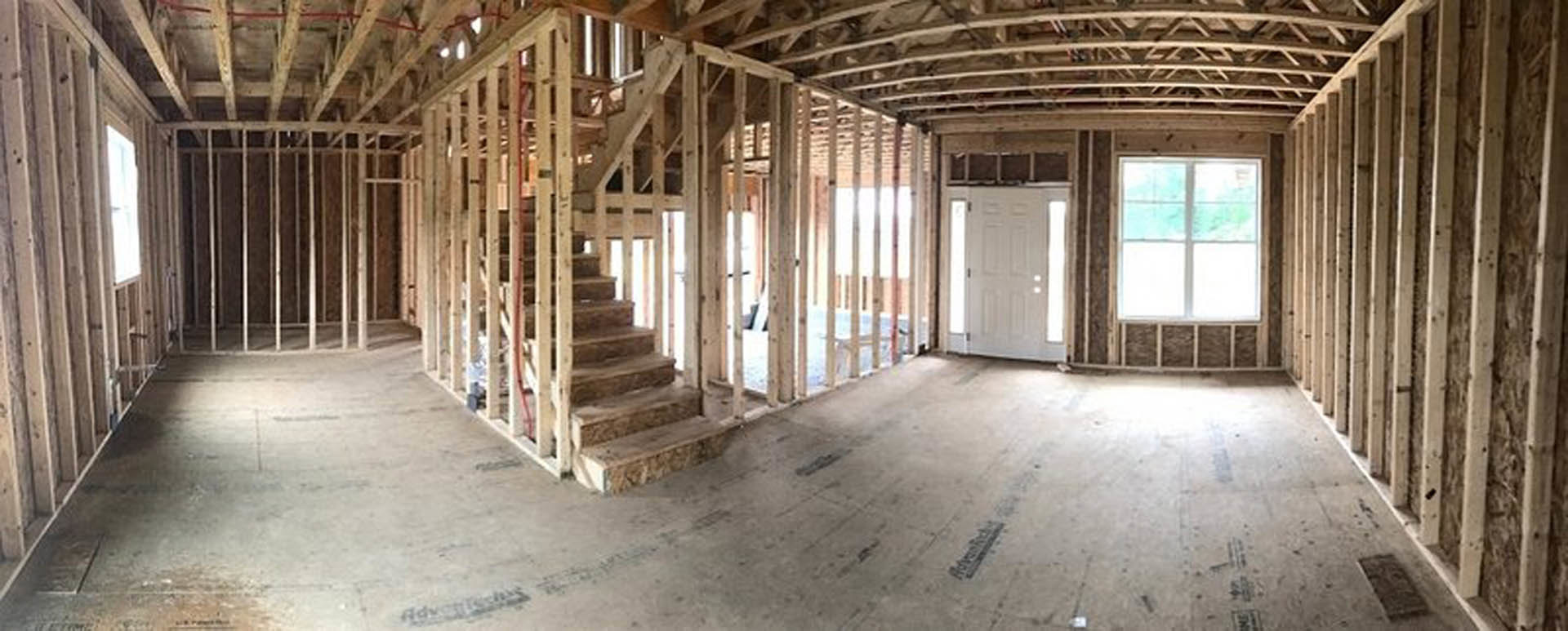 Unfinished interior with exposed wooden framing, white door illuminated by daylight, white-framed window, and partially built wooden staircase.