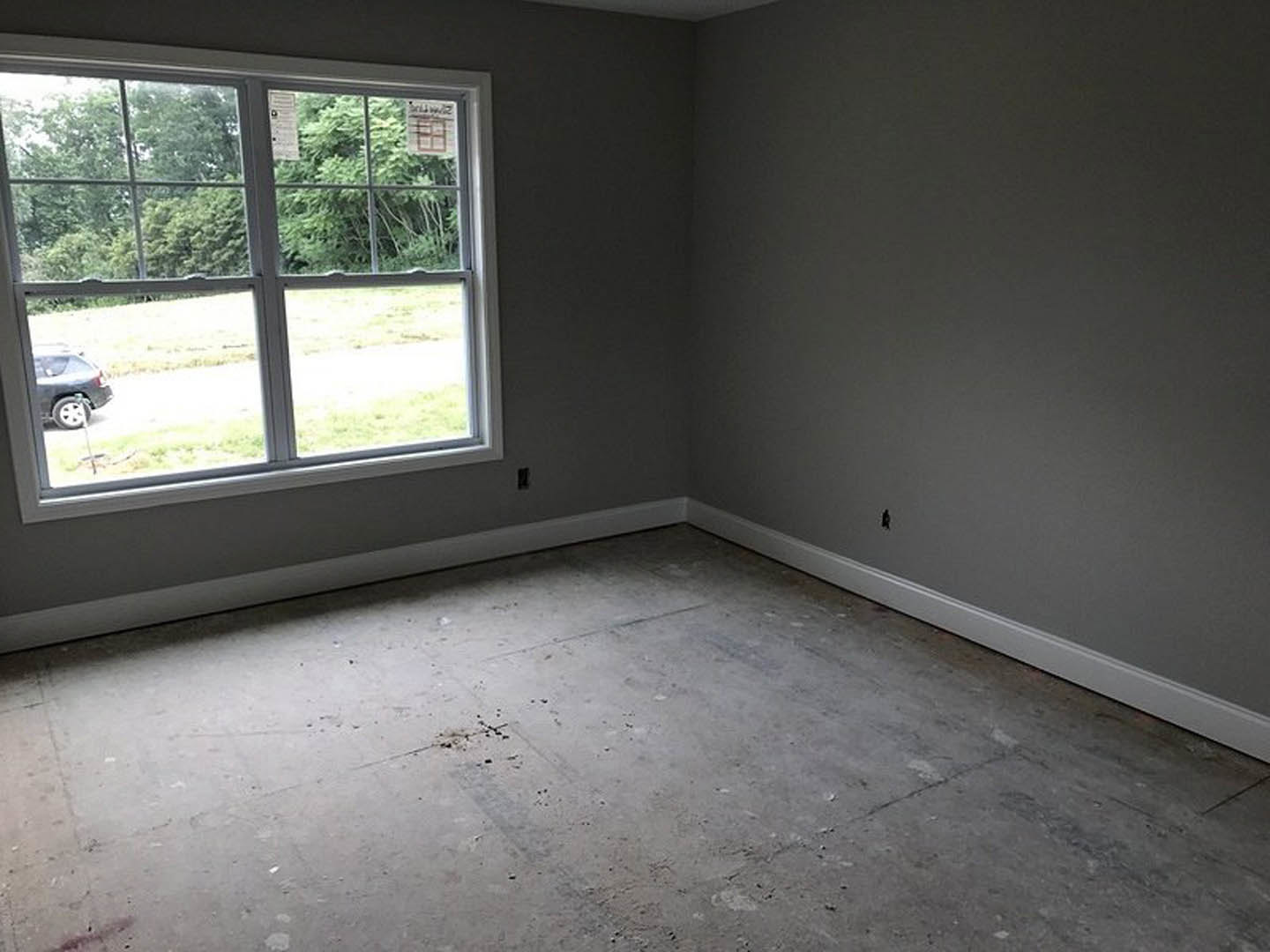 Concrete floor with white trim in a room featuring a large window, plaster walls, and daylight illuminating the interior; outside, a car is parked on grass beside the road.