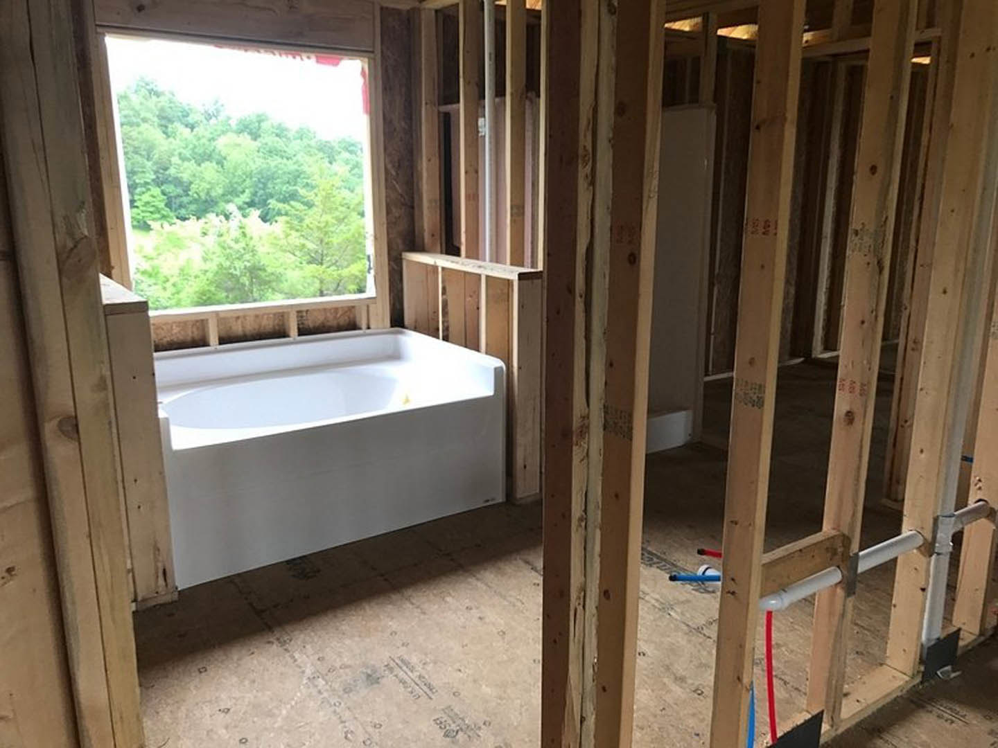 Bathroom under construction featuring exposed wood framing, white bathtub, window overlooking trees, and visible plumbing pipes.