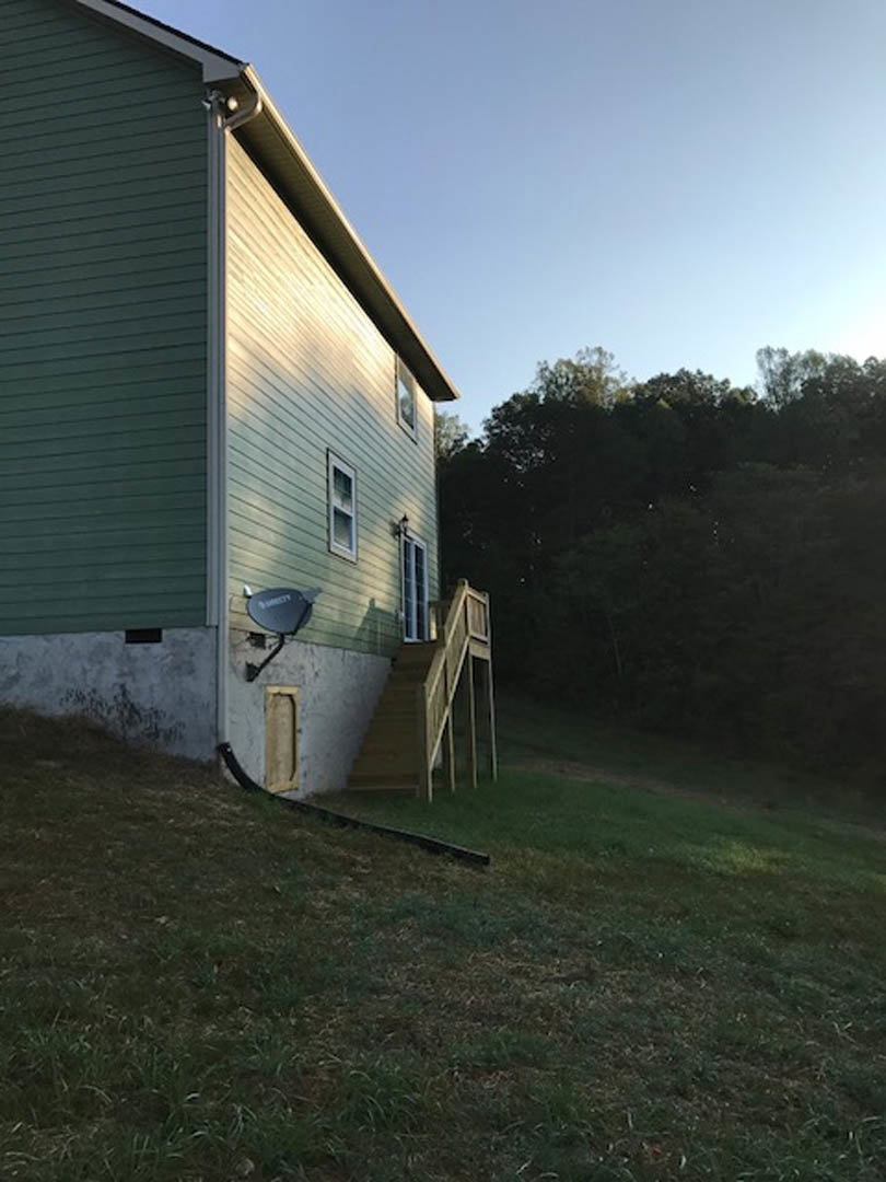 Modern two-story house with wood deck and staircase leading to backyard, surrounded by grass and trees, satellite dish mounted on side wall