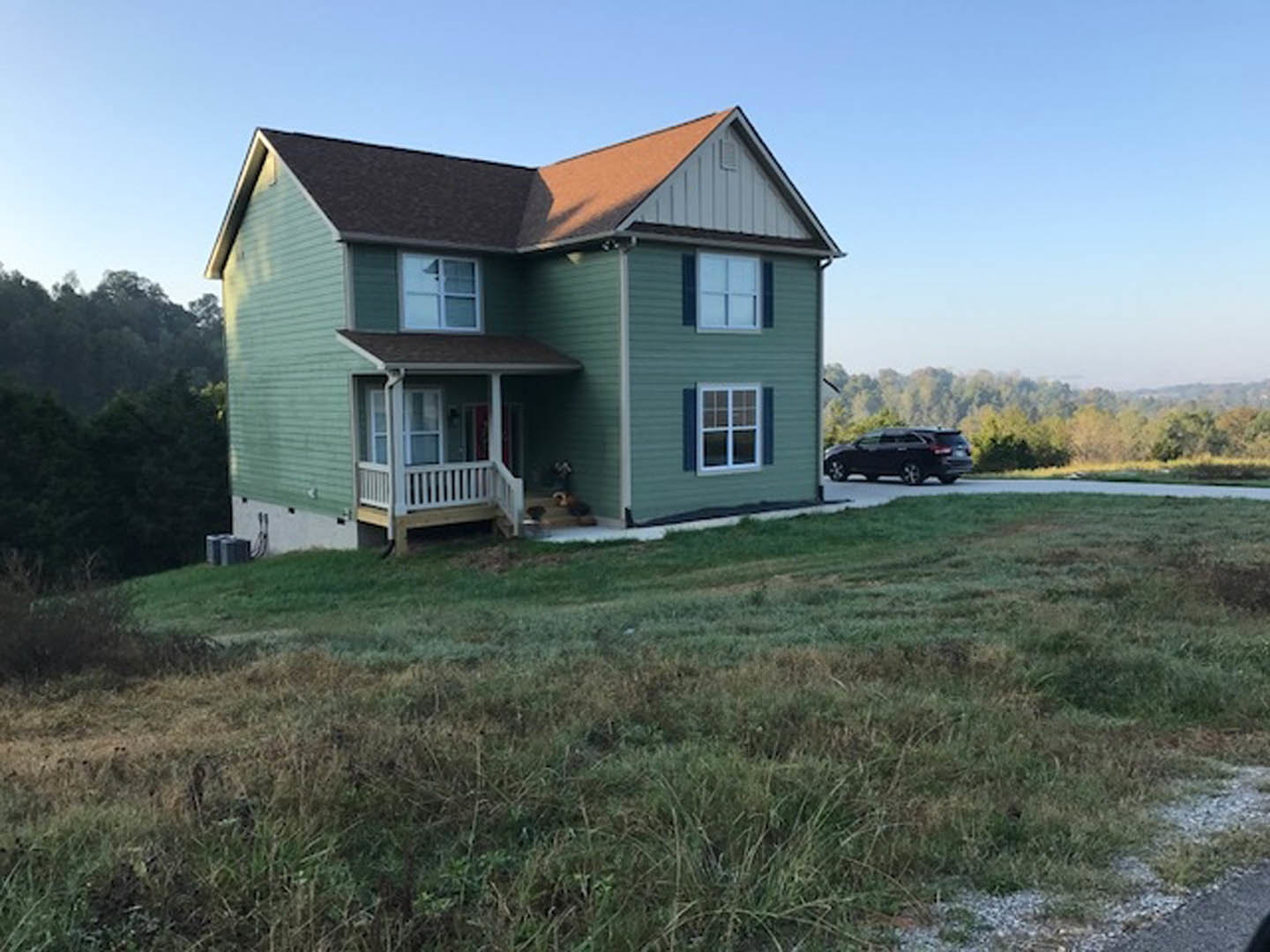 Green siding house with white porch, black car parked on driveway, row of square windows, grassy front yard, trees in background