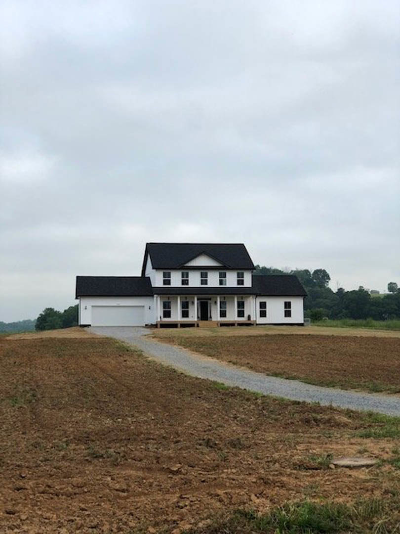 White house with black gabled roof, driveway connecting to dirt road, grassy lot bordered by distant trees under partly cloudy sky