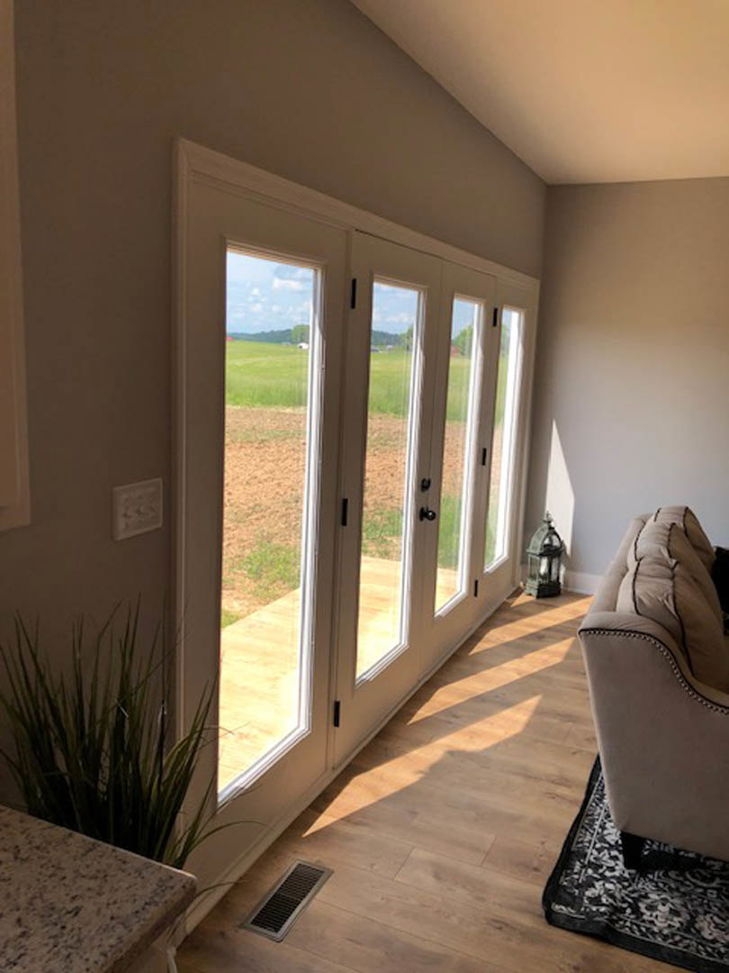 Living room with wide glass patio doors overlooking a grassy field, marble coffee table in foreground, upholstered chair beside a potted plant, light wood flooring with floor vent.