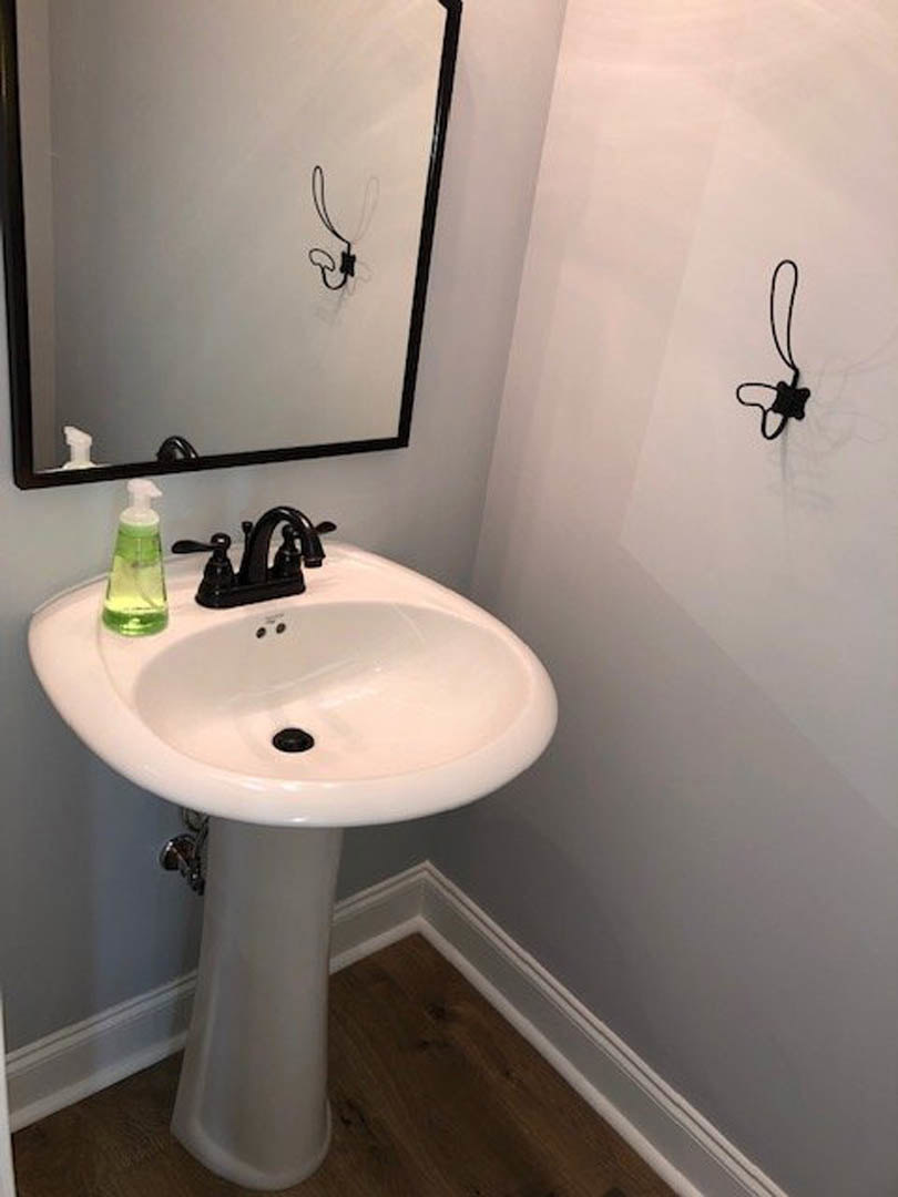 White bathroom sink with black faucet and handles, green bottle on countertop, rectangular mirror with black frame, light-colored wall and floor visible
