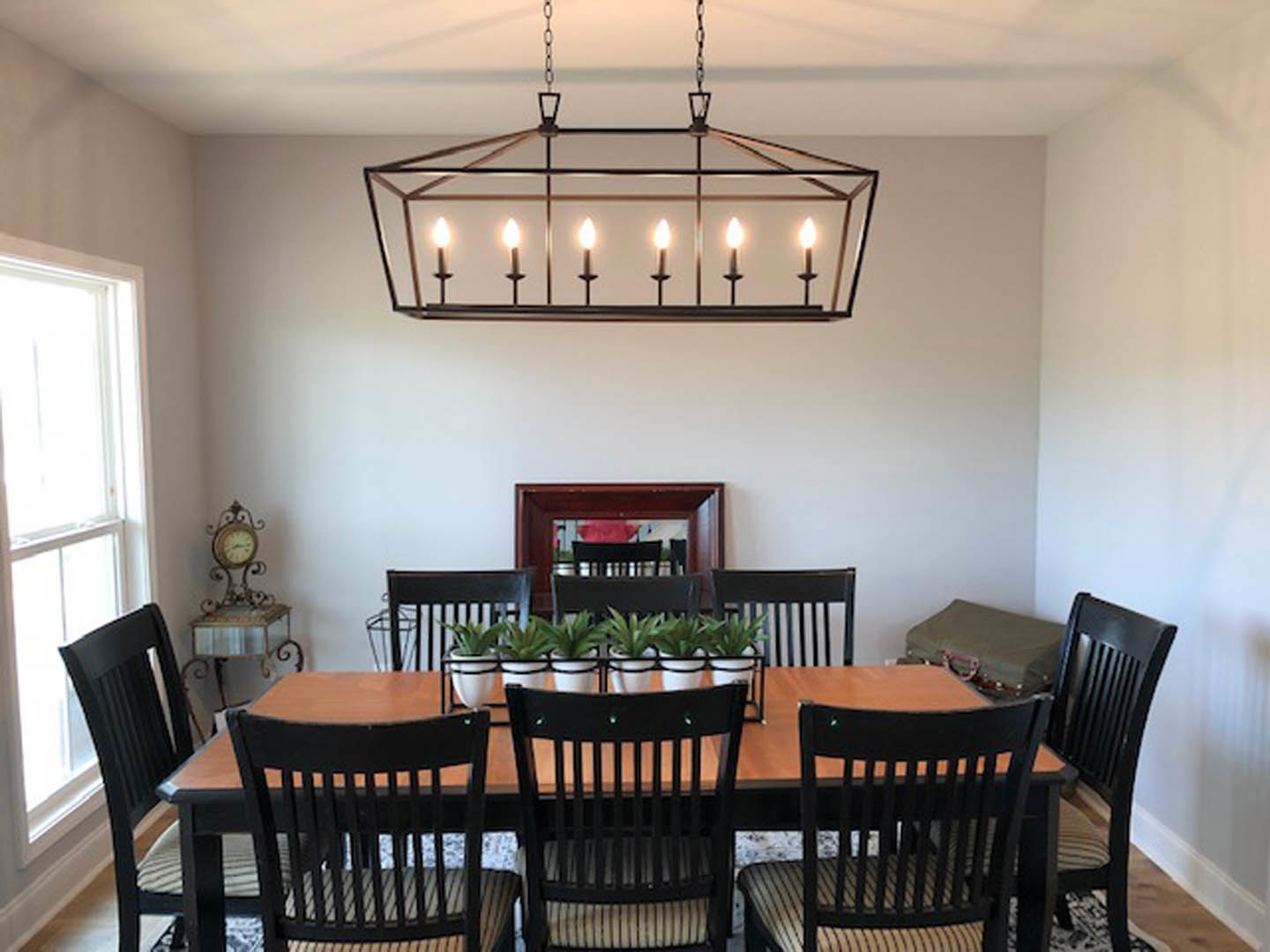 Wood dining table surrounded by upholstered chairs, glass chandelier overhead, neutral walls, decorative clock, potted plants, and metal-framed pendant lights.