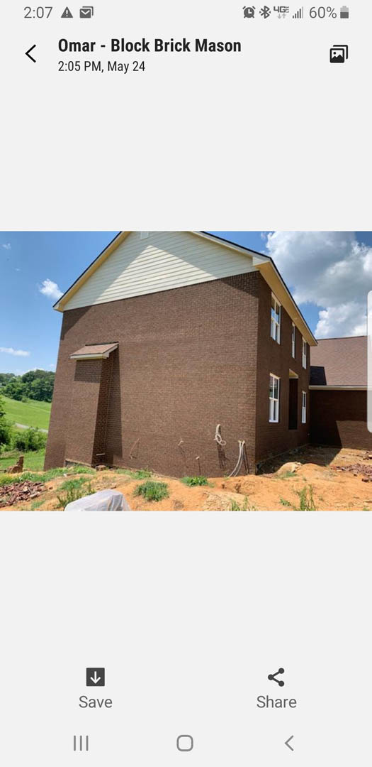 Partially built brown brick house with white roof, surrounded by grass and mature trees, under a cloudy sky