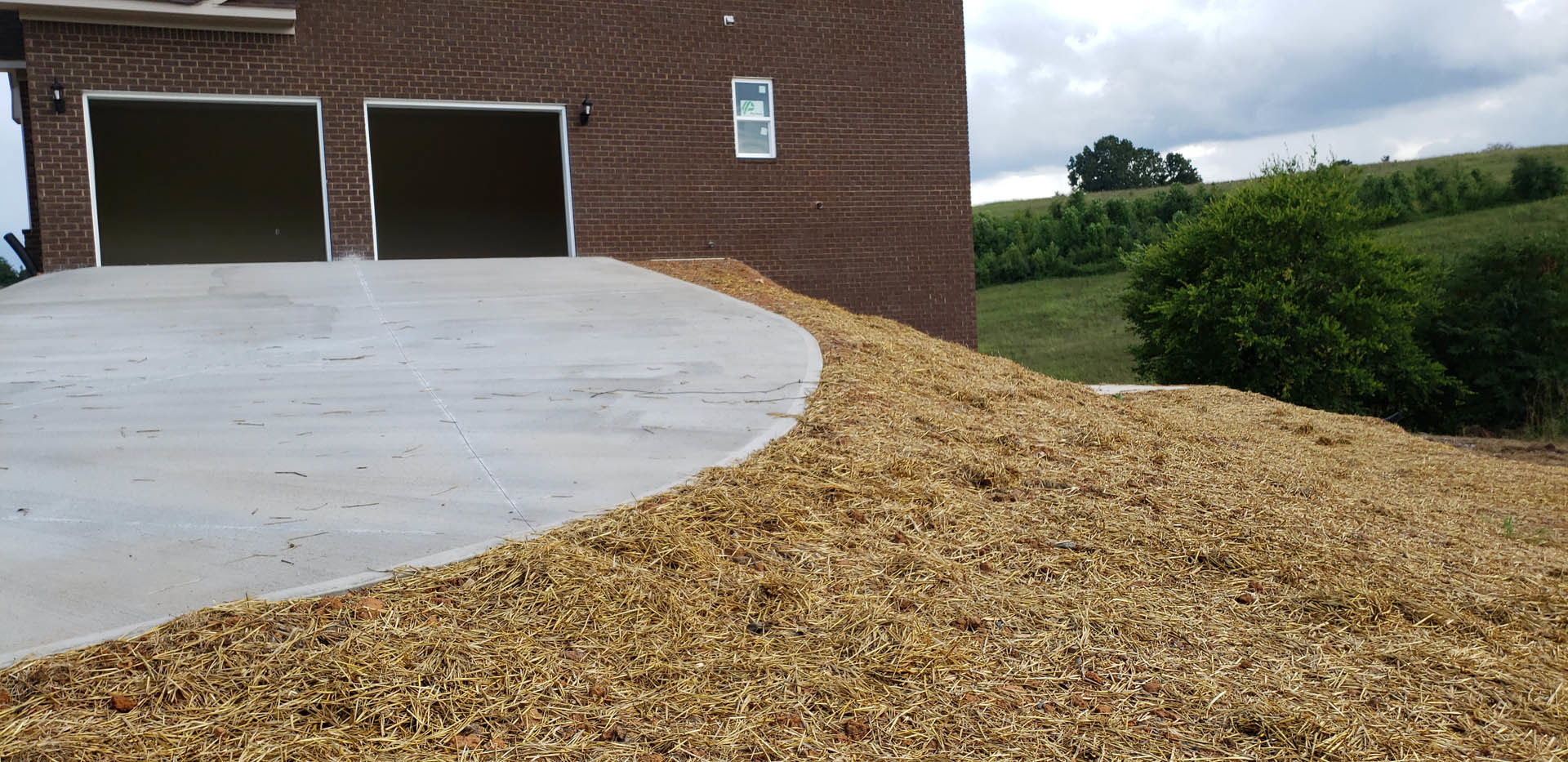 Concrete driveway with scattered straw, brick wall featuring a black screen, window displaying a sign, grassy area and trees in background under partly cloudy sky