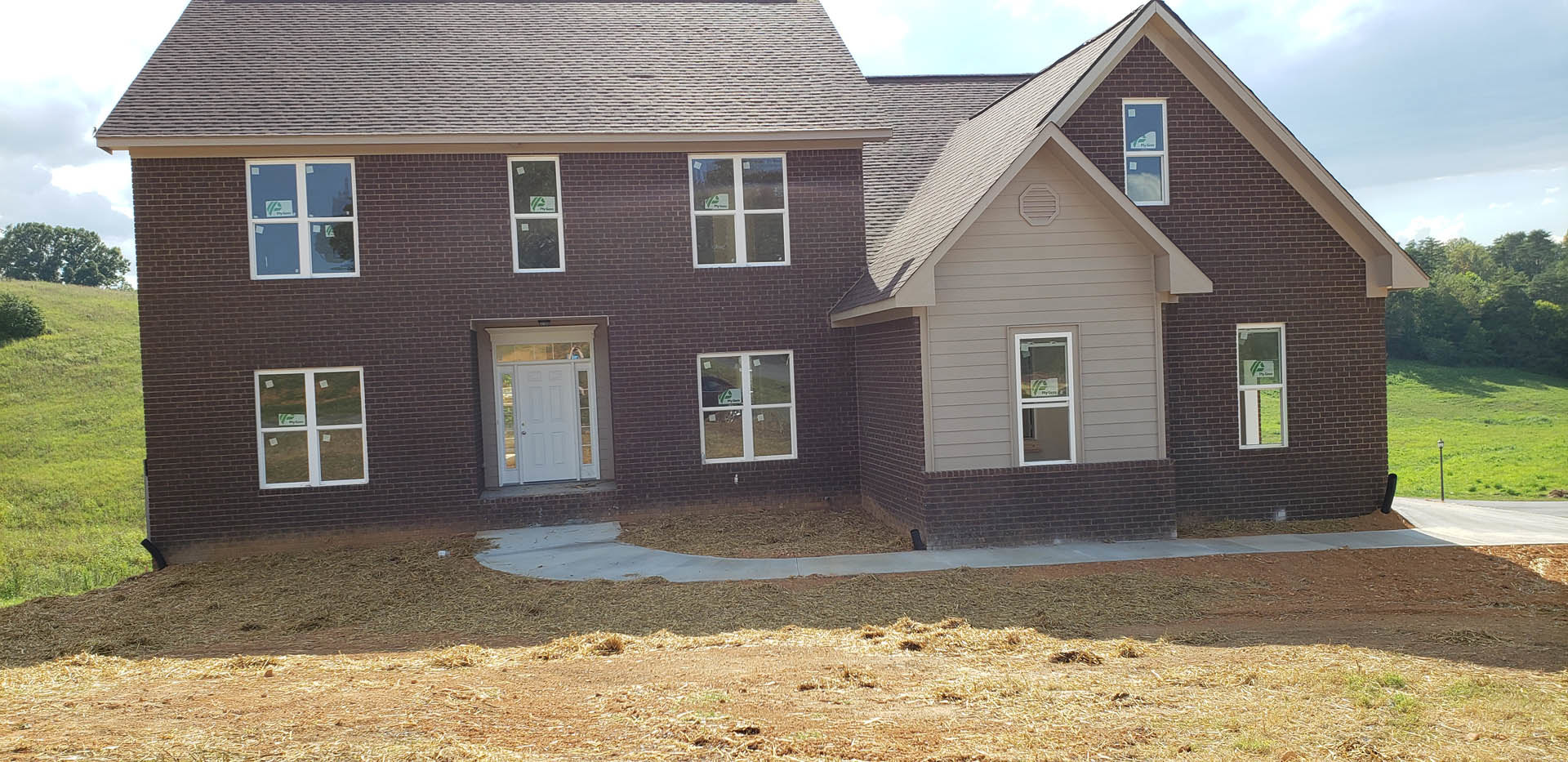 Two-story house with white siding, glass-paneled front door, multiple windows, dirt yard area with ramp, grassy lawn, and trees in background