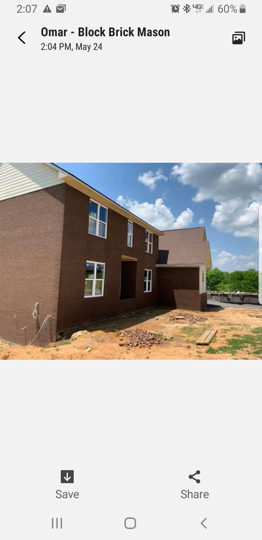 Partially built brick house with exposed framing, several installed windows, and construction materials scattered in front; clear blue sky and distant trees in background