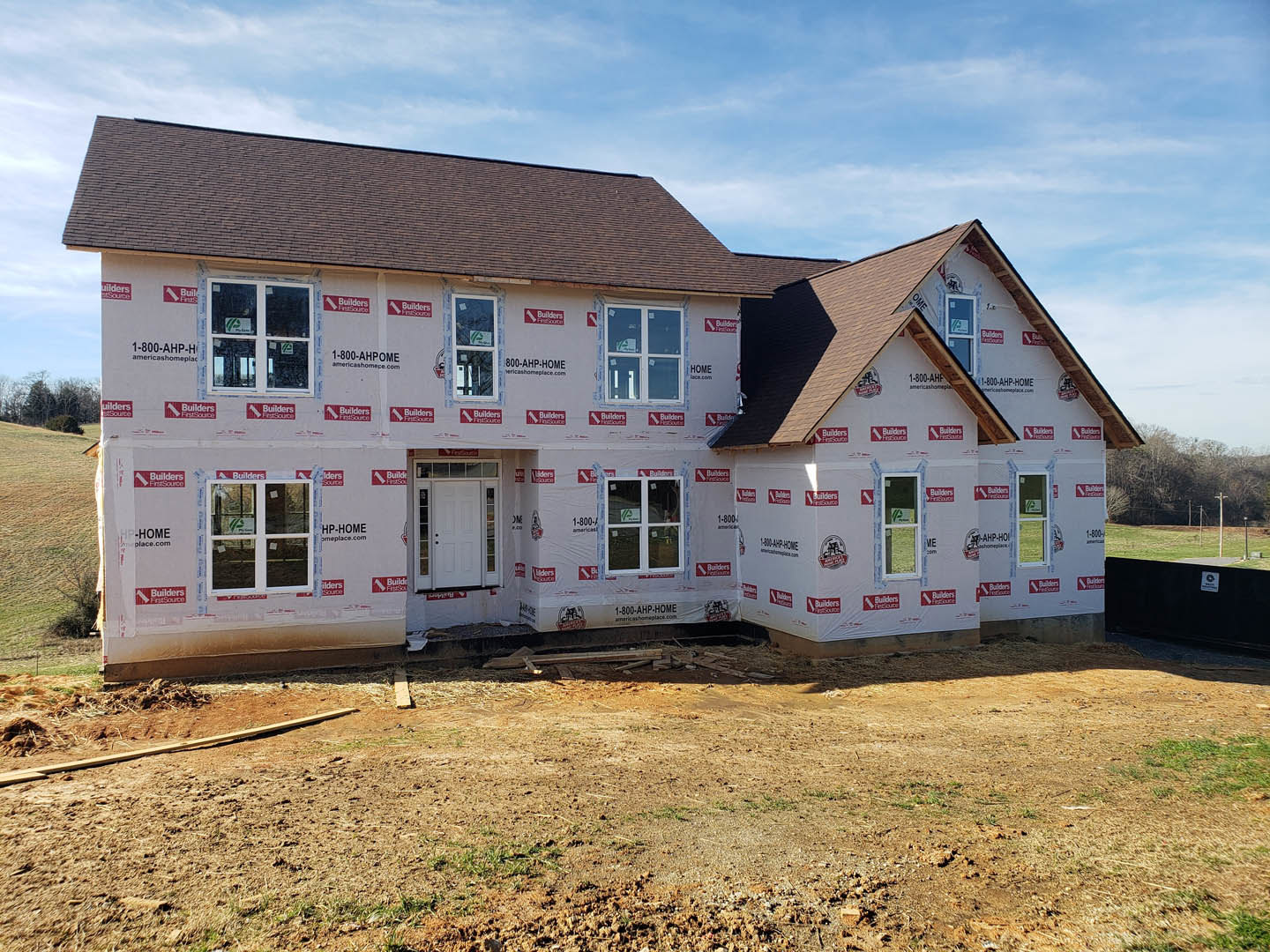 Partially built house with white and red insulation wrap, glass paneled door, multiple windows including one with a sign, dirt yard, and cloudy sky