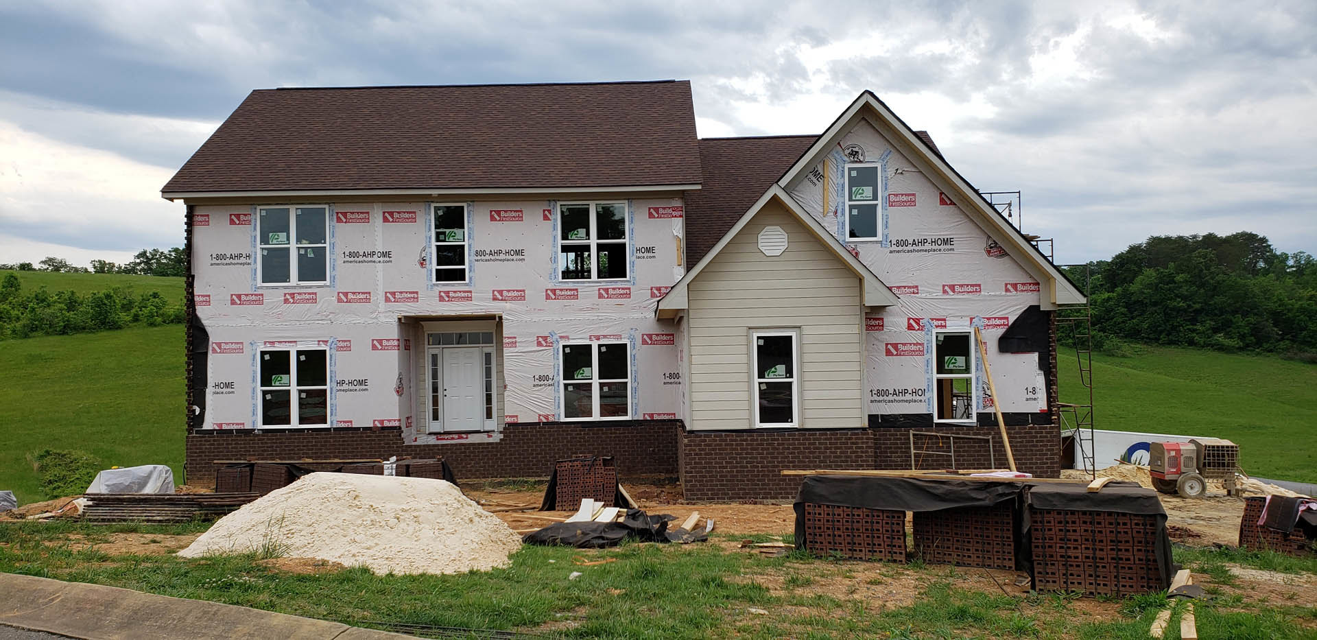 Two-story house under construction with white door and windows, large pile of white sand in front, stack of bricks on site, white vent on wall, sign taped to window, grassy yard