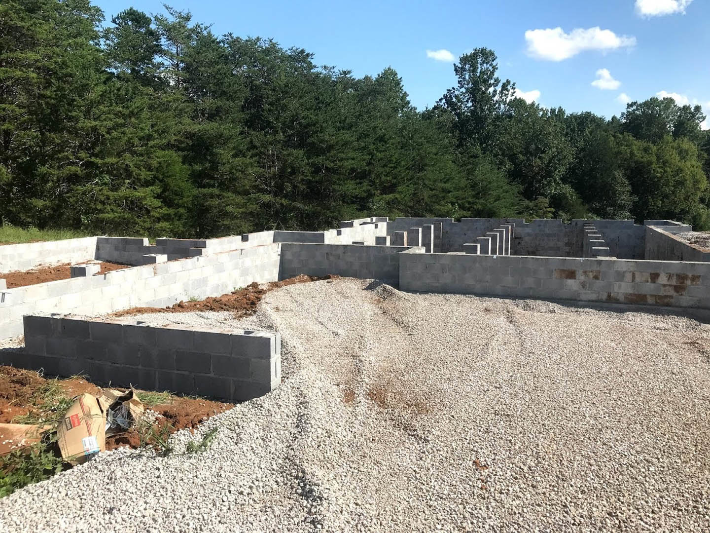 Gravel pile and bricks on a construction site bordered by concrete block wall, surrounded by trees under cloudy sky