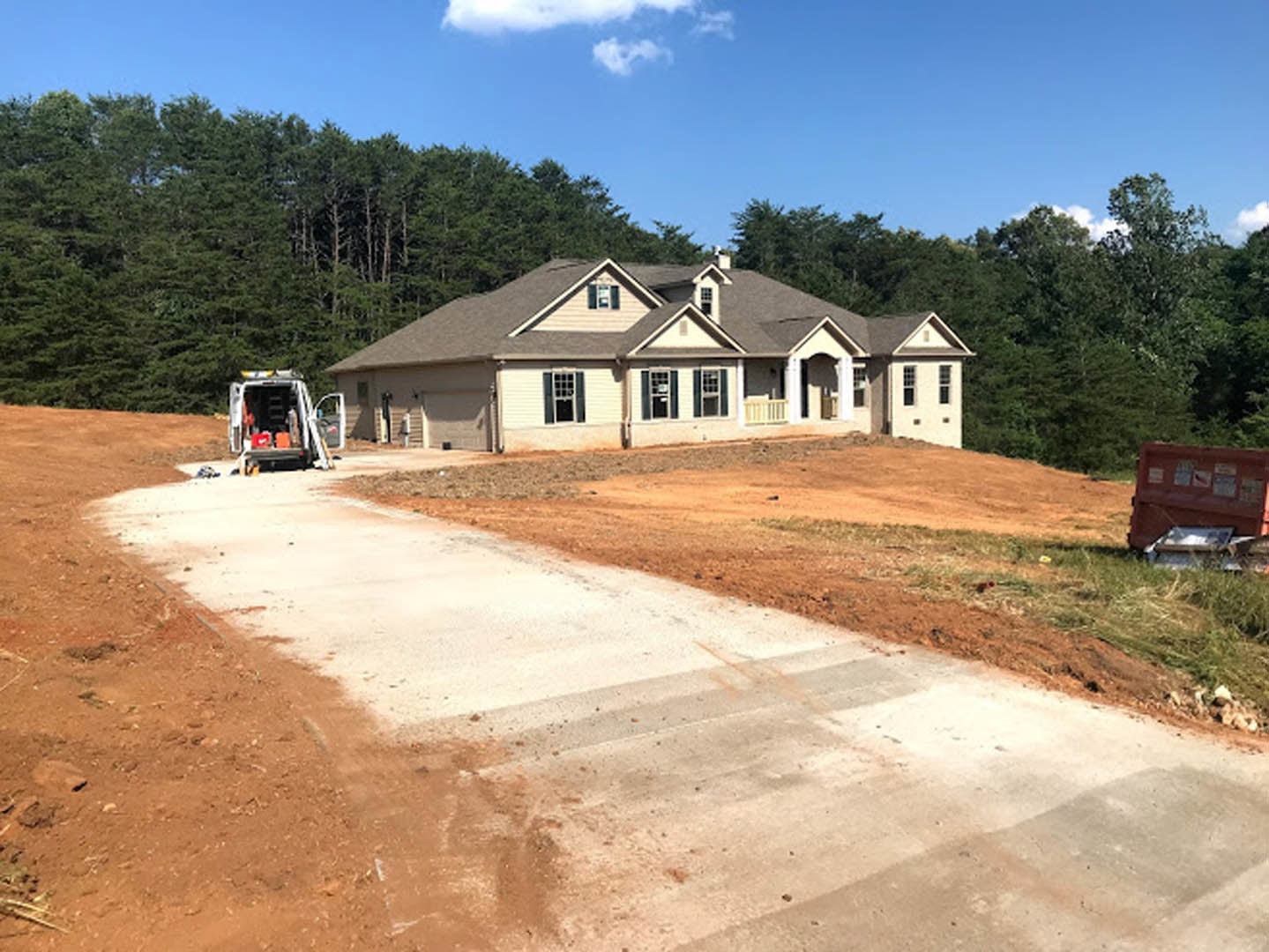 Two-story house with a few windows, driveway leading to a dirt road, white van with open doors parked beside a metal container with signage, blue sky with scattered clouds, trees