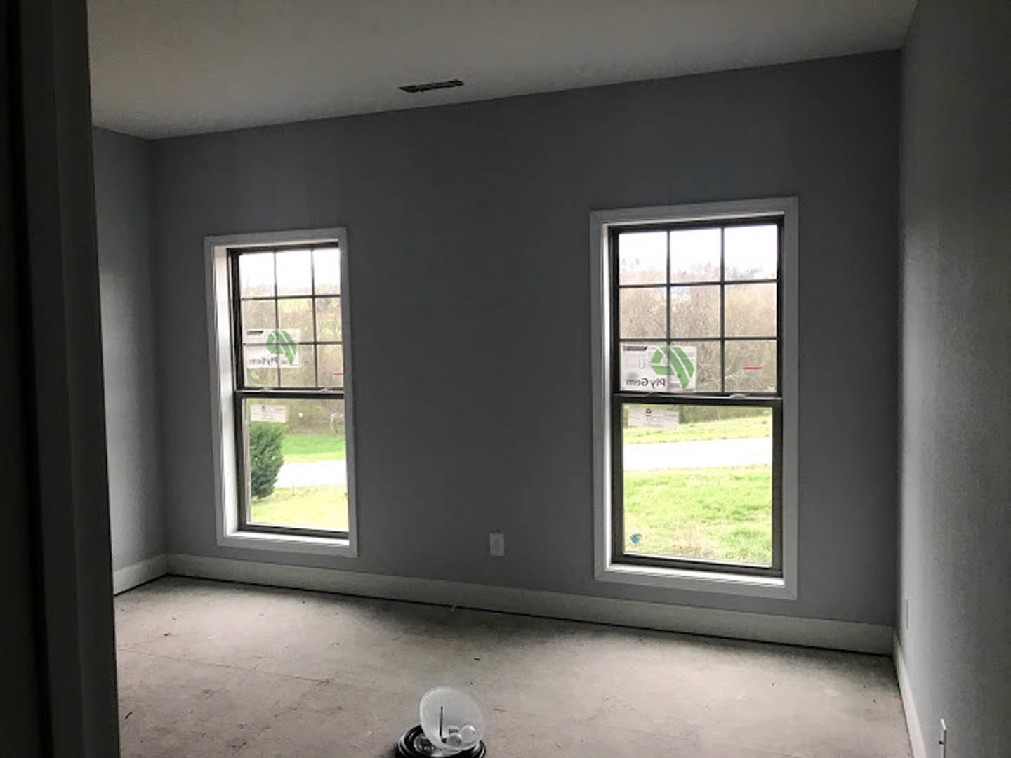 Modern room with two large windows, concrete floor, plaster walls, window blinds, and a ceiling light fixture; sunlight streams in, highlighting minimalist finishes.