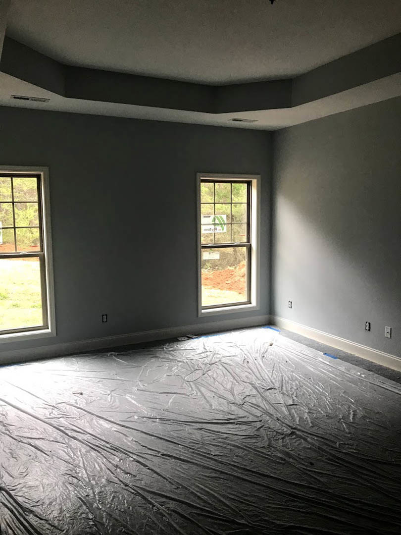 Unfinished room with plastic-covered floor, two windows—one displaying a sign and one overlooking a grassy field—white plaster ceiling, and sunlight streaming in