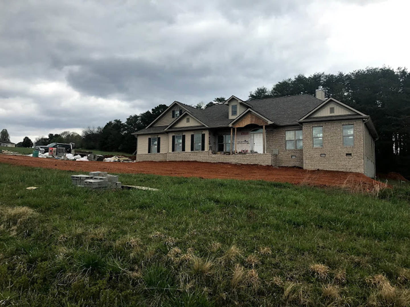 Partially built house with exposed framing, surrounded by green grass and mature trees, under a cloudy sky