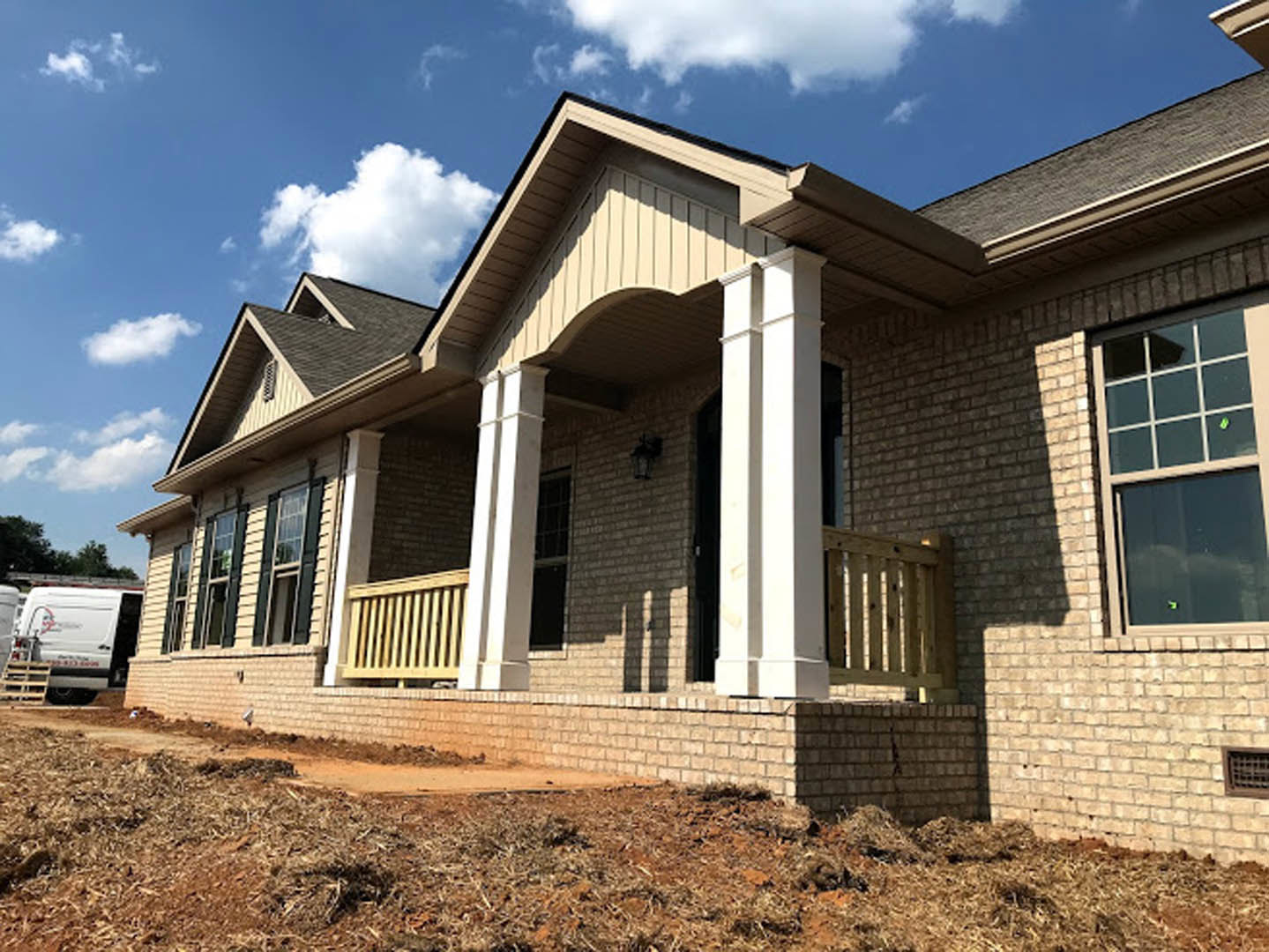 Partially built house with exposed wooden framing, white siding, covered porch, white pillars, wooden fence, white van parked nearby, green leaf sticker on window, blue sky with