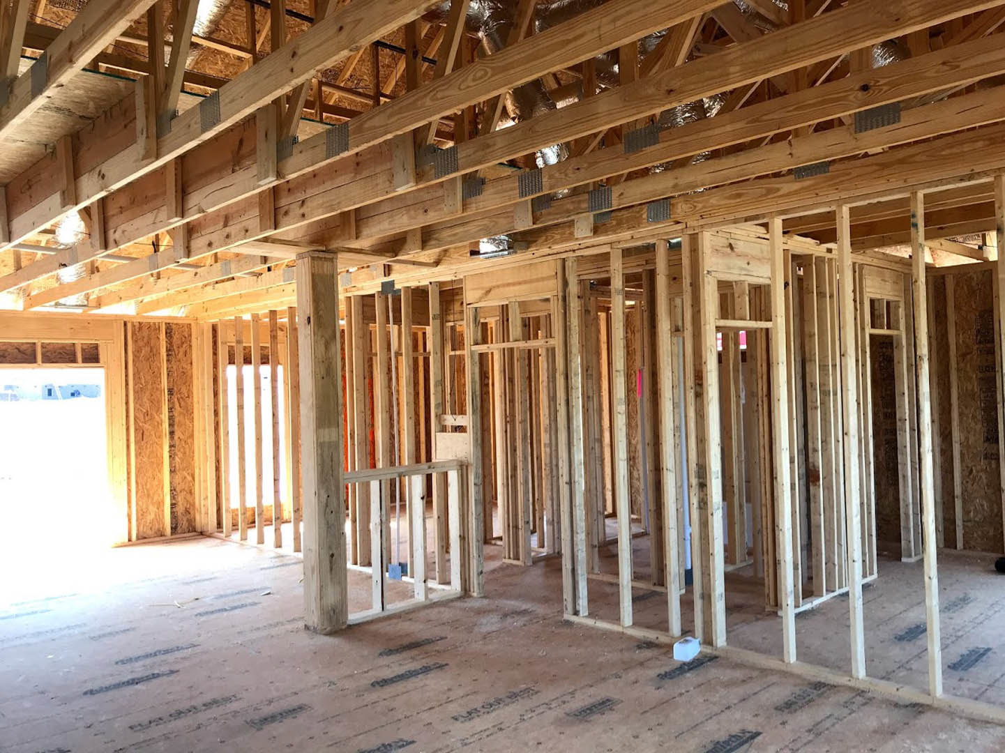 Exposed wooden beams and framing inside a custom home under construction, unfinished ceiling and floor visible, bright lighting highlighting lumber structure