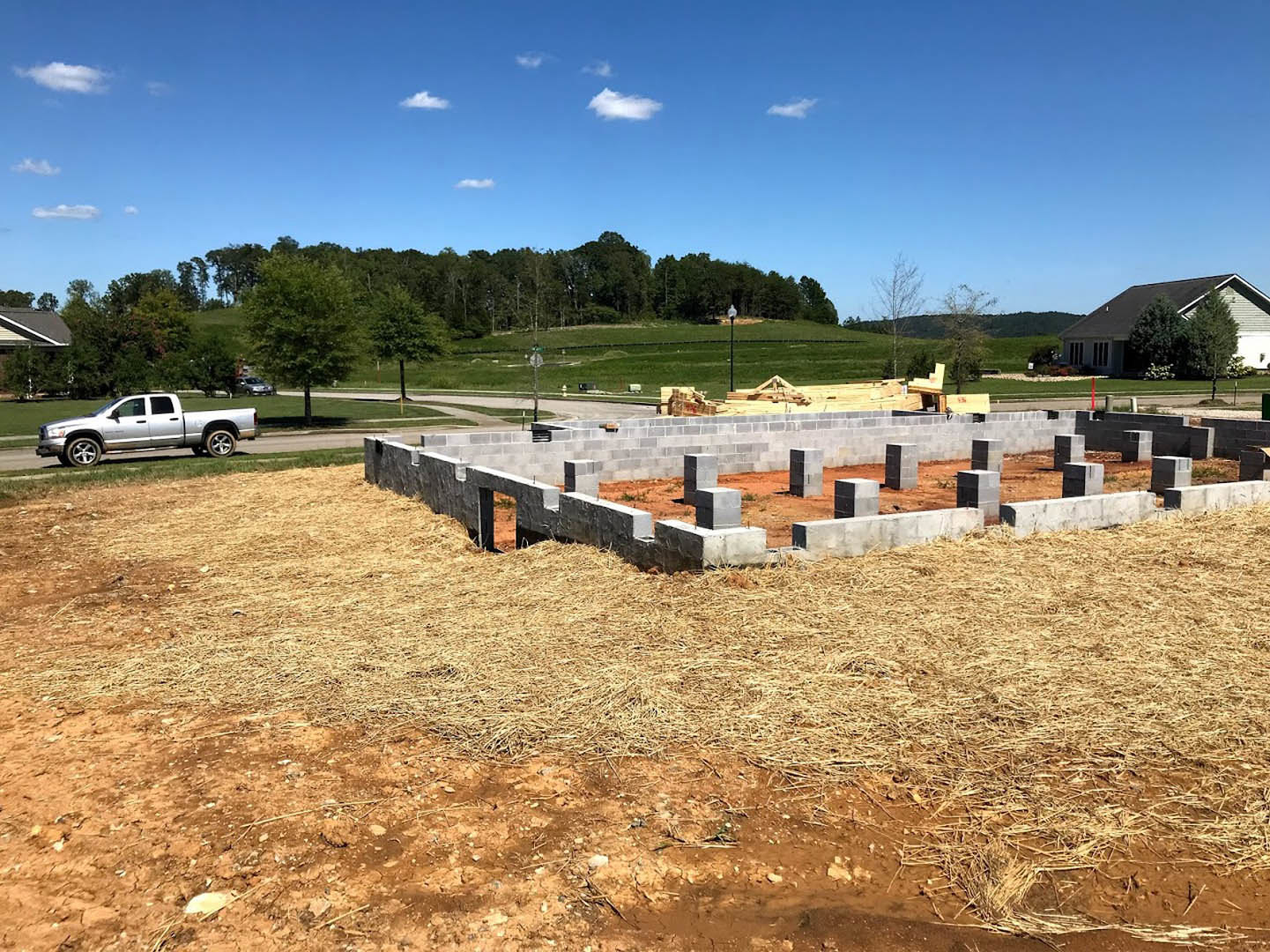 Framed house under construction with exposed wooden beams, stacks of concrete blocks, white pickup truck parked nearby, dirt and grass foreground, leafy trees and cloudy sky in
