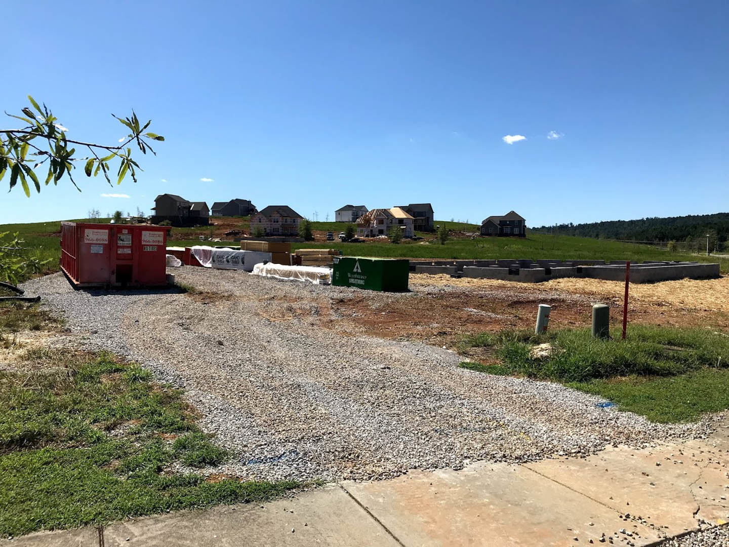 Gravel driveway leading to a distant building, red container with signage, green utility box with white text, tree branch in foreground, blue sky with scattered clouds above