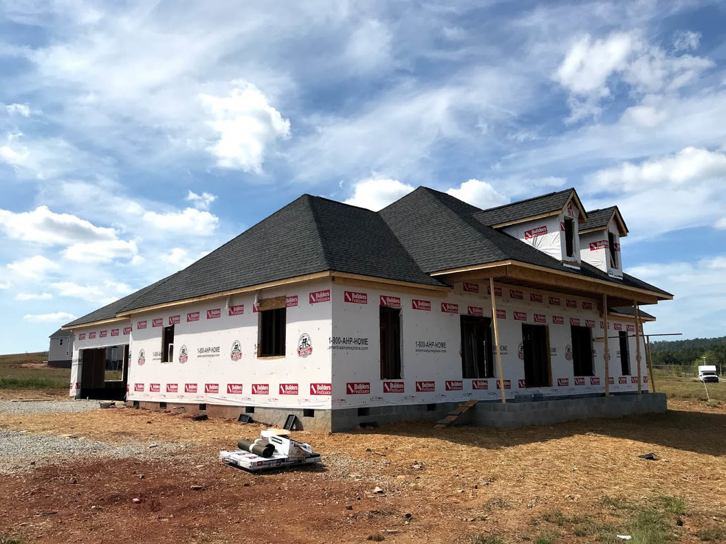 Partially built house with exposed framing, red tape along exterior wall, rolls of black material on white box, red and white signs, grassy yard, blue sky with clouds