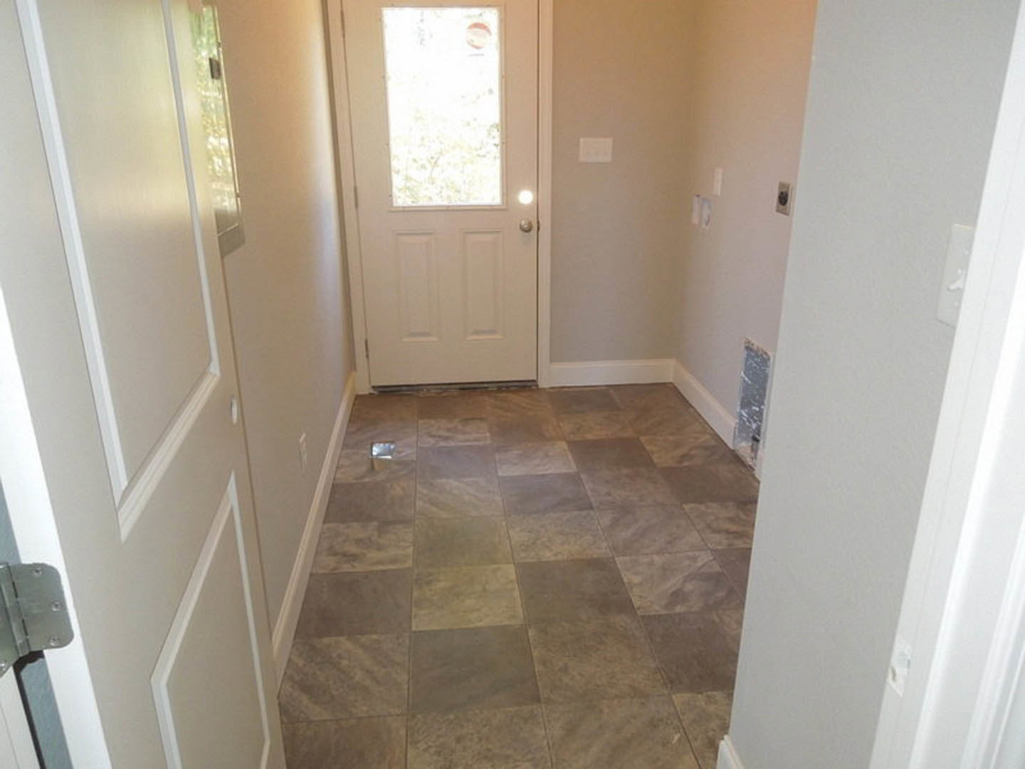 Hallway with light-colored tile flooring featuring a hole, white door with window and white-framed window, light switch on wall, plaster walls, partial blurry face visible