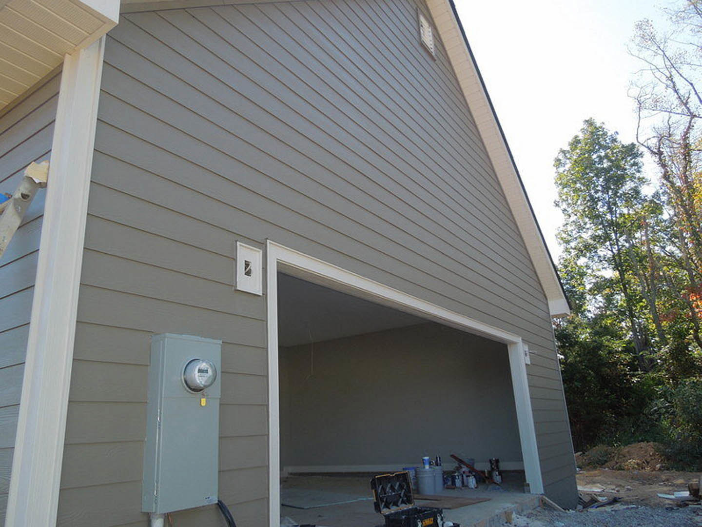 Open garage with white door, grey siding, window on white wall, leafy tree nearby, close-up of utility meter, blue sky overhead