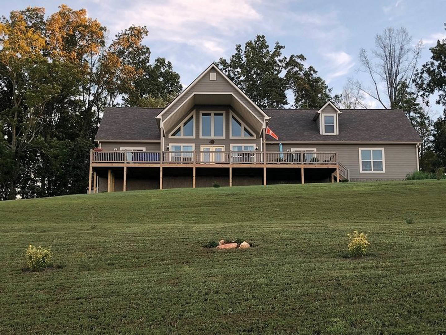 Two-story farmhouse with white siding, red triangle flag on deck, white-framed windows, spacious porch with railing, large green lawn, scattered trees, partly cloudy sky