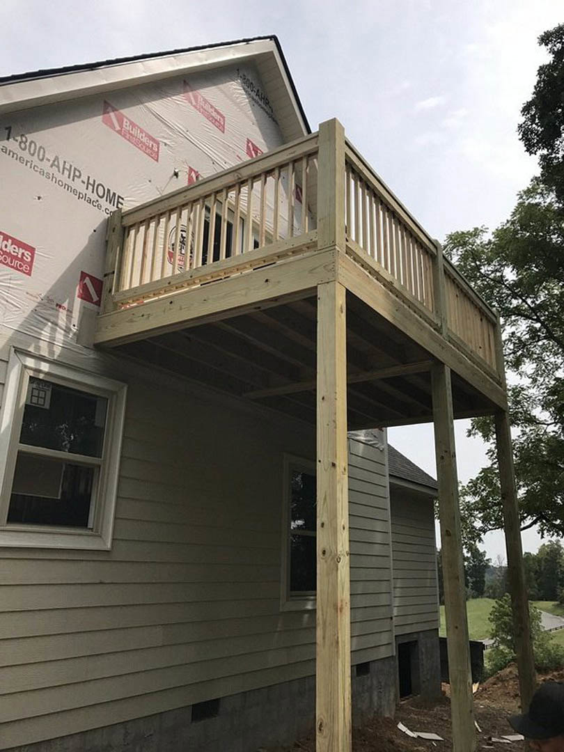Wooden deck with white railing attached to house, person in black hat standing near red and white sign, window with broken glass, trees and sky in background