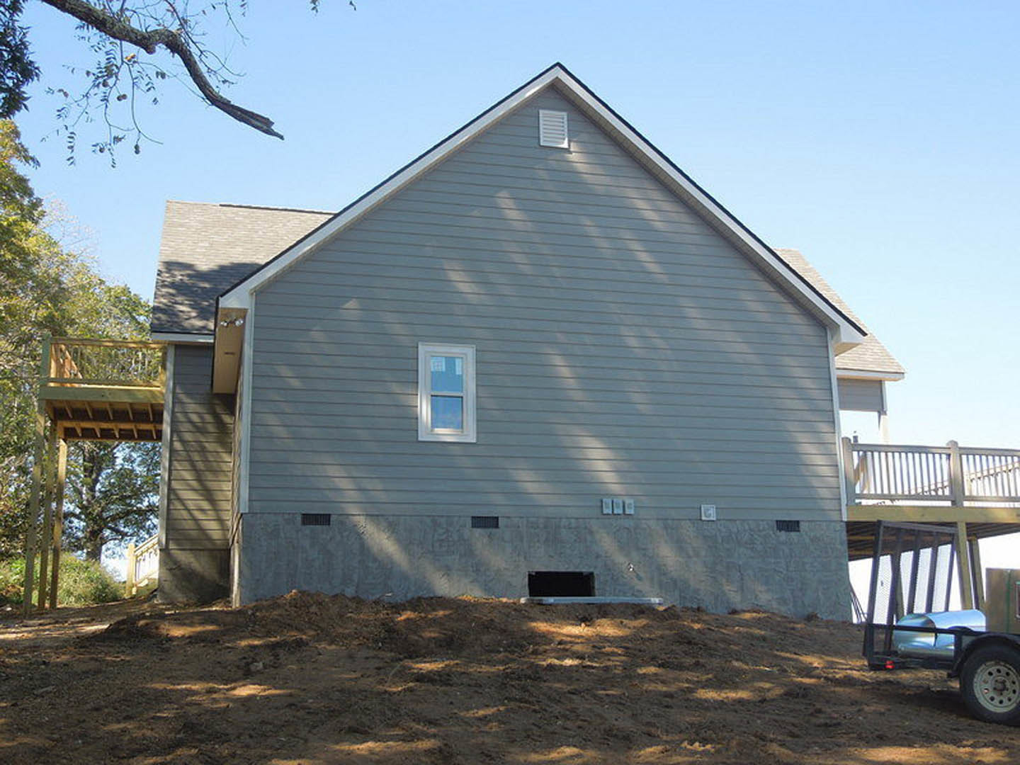 Wood-framed house under construction with exposed siding, partially built deck, large window, roof vent, dirt yard, and a person standing near the foundation.