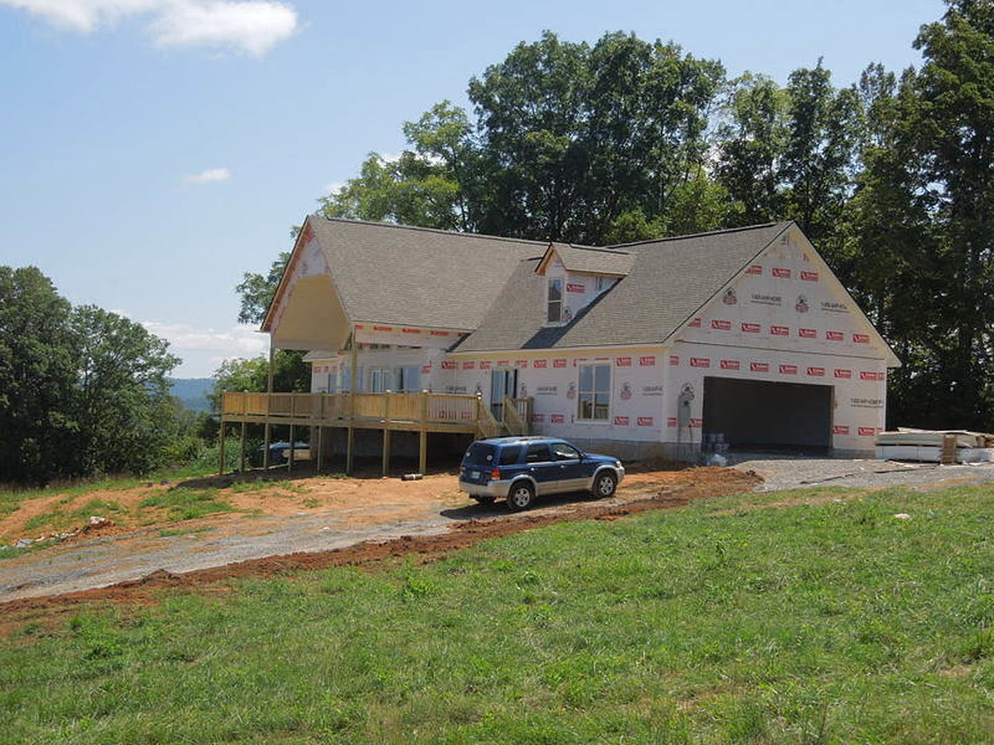 Two-story house with attached garage, wooden deck with railing, blue SUV parked on dirt driveway, red and white tape along side of house, grassy yard, trees, and partly cloudy sky