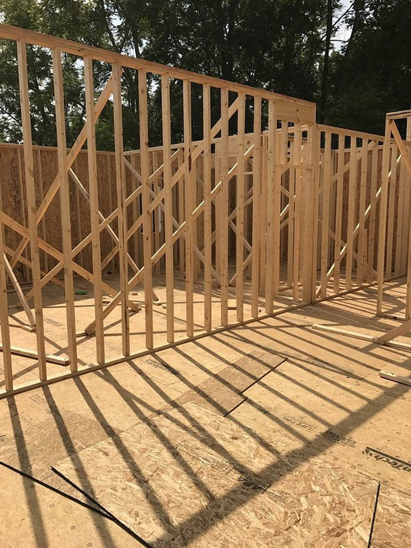 Wood-framed house under construction with exposed lumber, wooden railing, and tree-lined background; shadows from a nearby fence cast across the unfinished floor and wall.