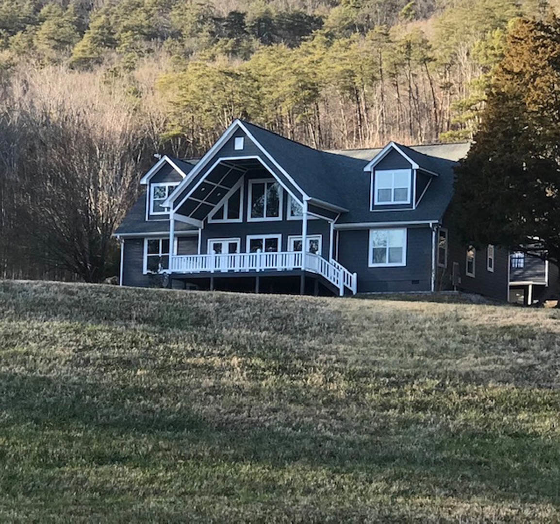 Spacious porch with white railing, wide stairs leading to grassy lawn, large leafy tree behind farmhouse-style home, multiple windows with white frames