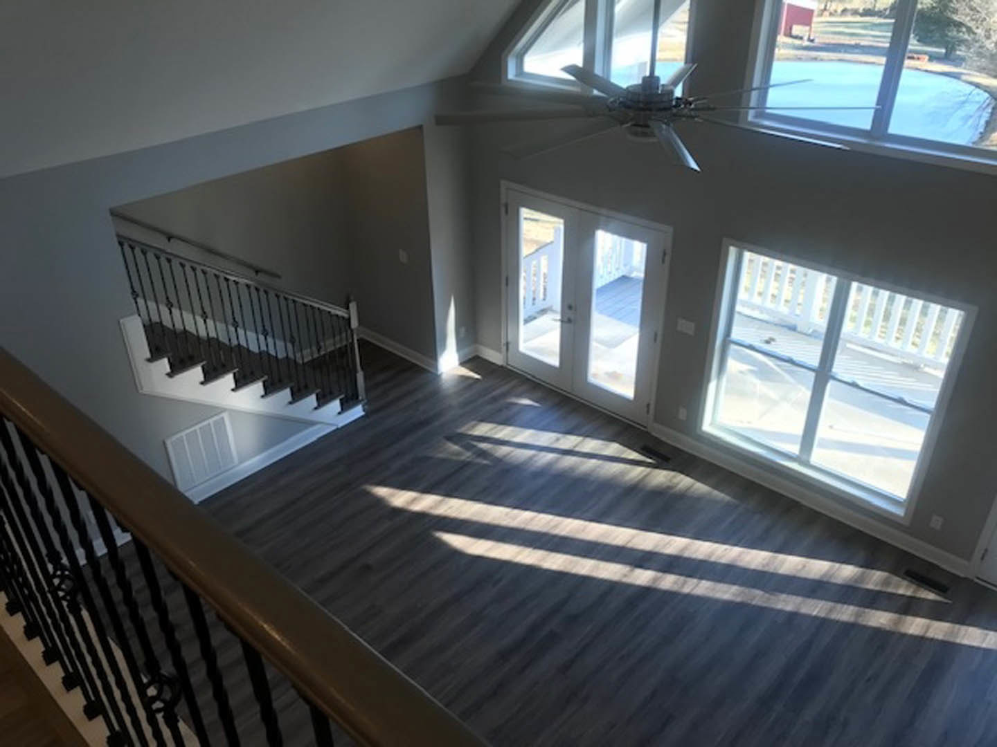 Wood-floored room featuring a ceiling fan, large windows, double glass-paneled doors, and a staircase with metal railings.