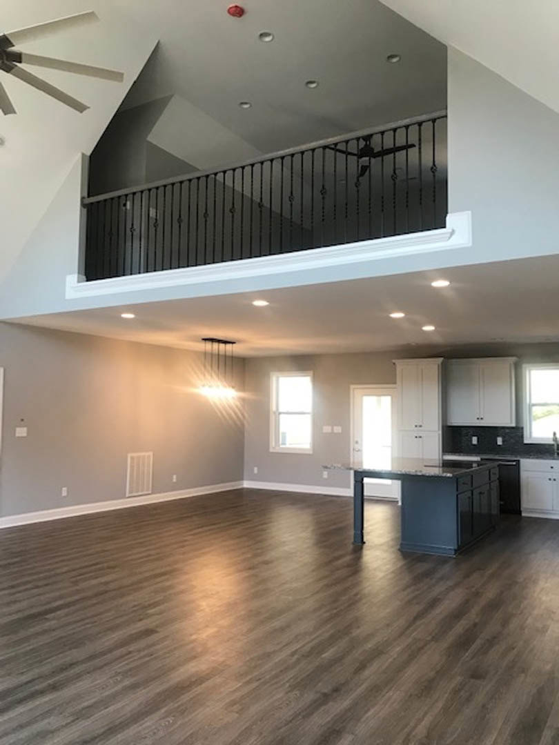 Open-concept room featuring a black kitchen island with drawers, hardwood flooring, white staircase with black railing leading to loft, white door with wall sconce, and window with