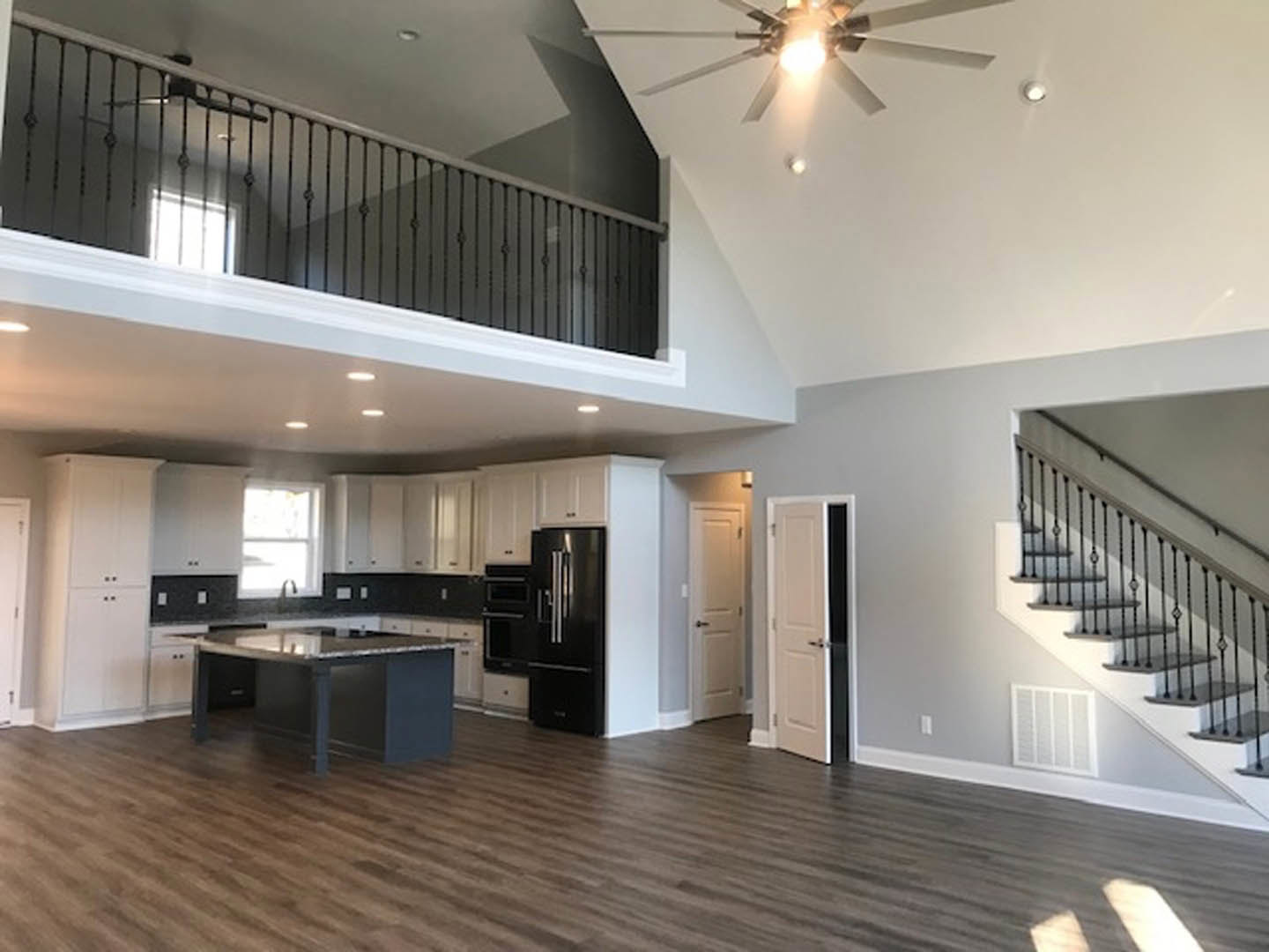 Open-concept room featuring a modern kitchen with white cabinetry, black accents, a ceiling fan with light, black metal stair railing, and a black door with metal handle against
