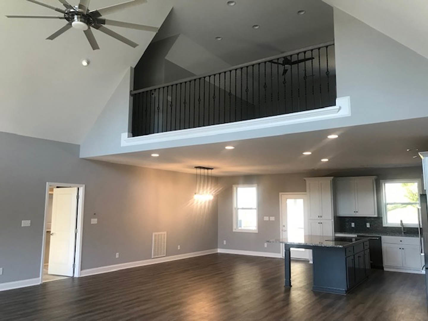 Open kitchen with marble-topped island, wooden dining table, ceiling fan with light, white walls, black railing, and window with white frame