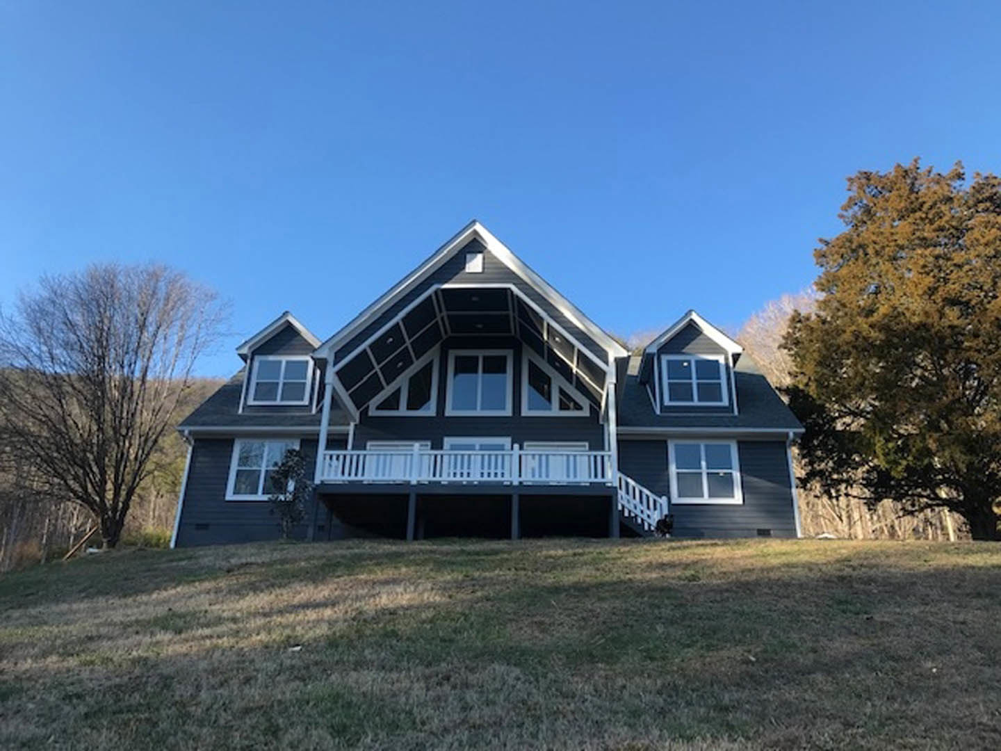 Two-story house with white siding, covered front porch, upper balcony with white railing, wooden deck, large windows, green lawn, mature trees with and without leaves.