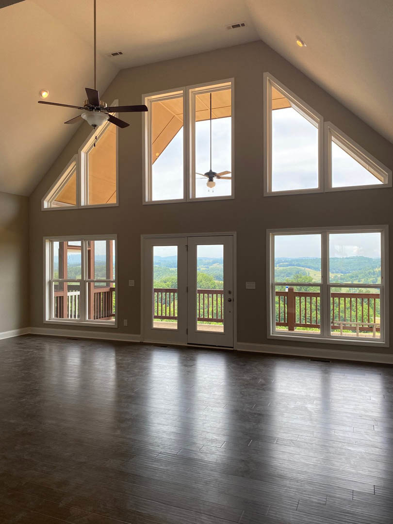 Spacious living area with dark wood flooring, white walls, ceiling fan with light fixture, multiple large windows, and double glass-paneled doors
