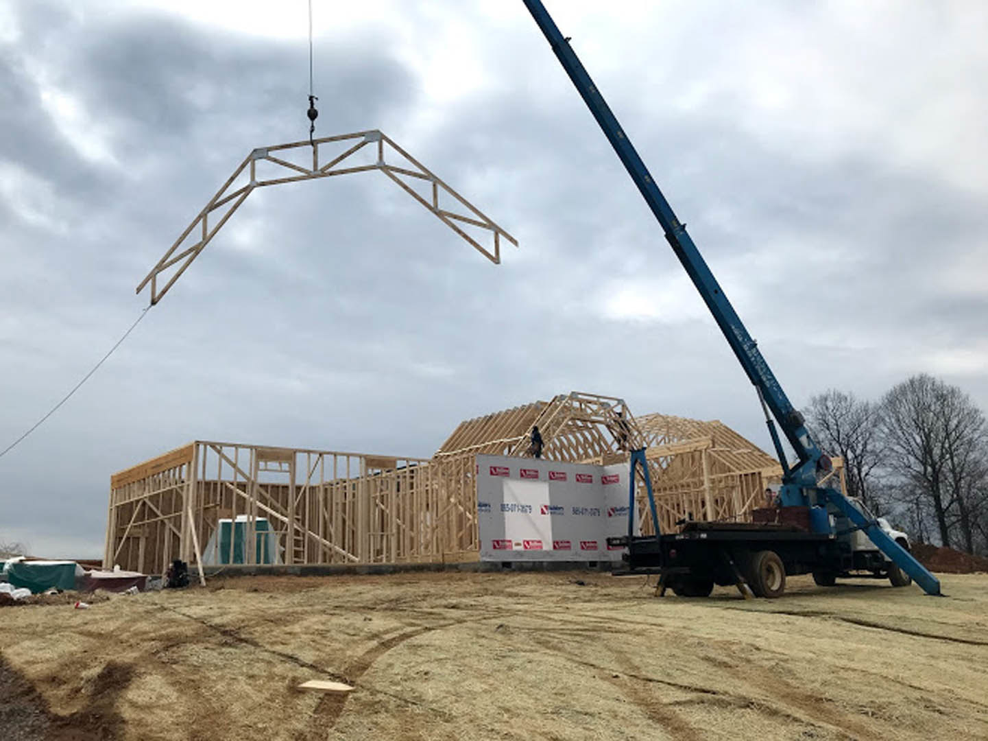 Crane hoisting a metal beam beside a partially constructed custom home, with construction vehicles and equipment on dirt ground under a cloudy sky