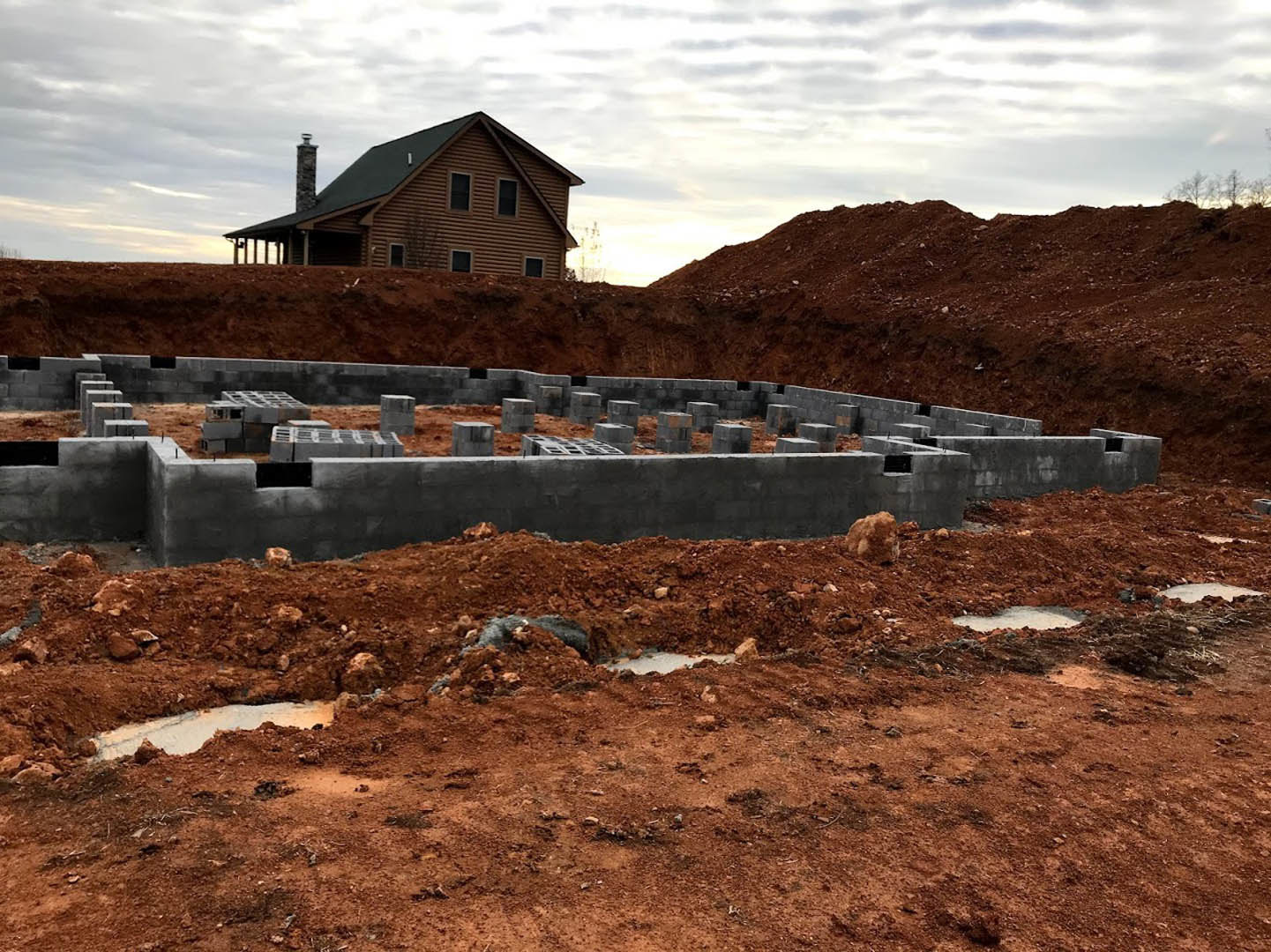 Wood-sided custom home with stone chimney, concrete foundation, and dirt landscaping under a cloudy sky