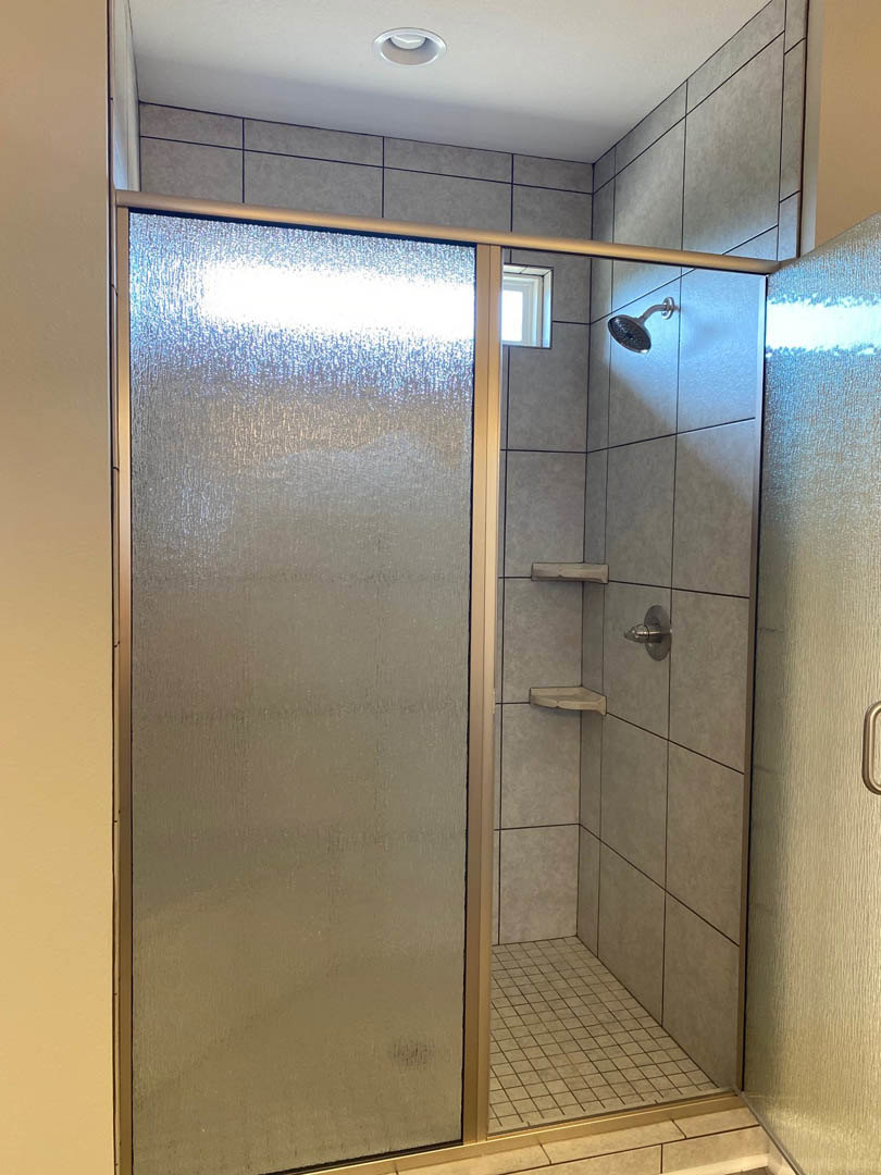 Walk-in shower with glass door and metal frame, wall-mounted shower head, ceiling light fixture, window letting in natural light, close-up of tiled floor