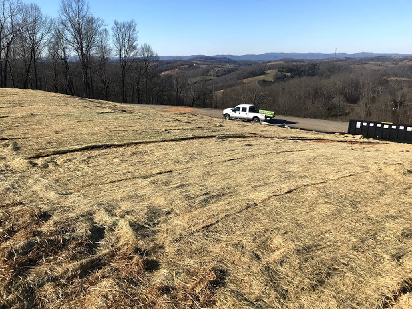 White pickup truck parked on a paved road atop grassy hill, surrounded by trees and bushes under clear blue sky