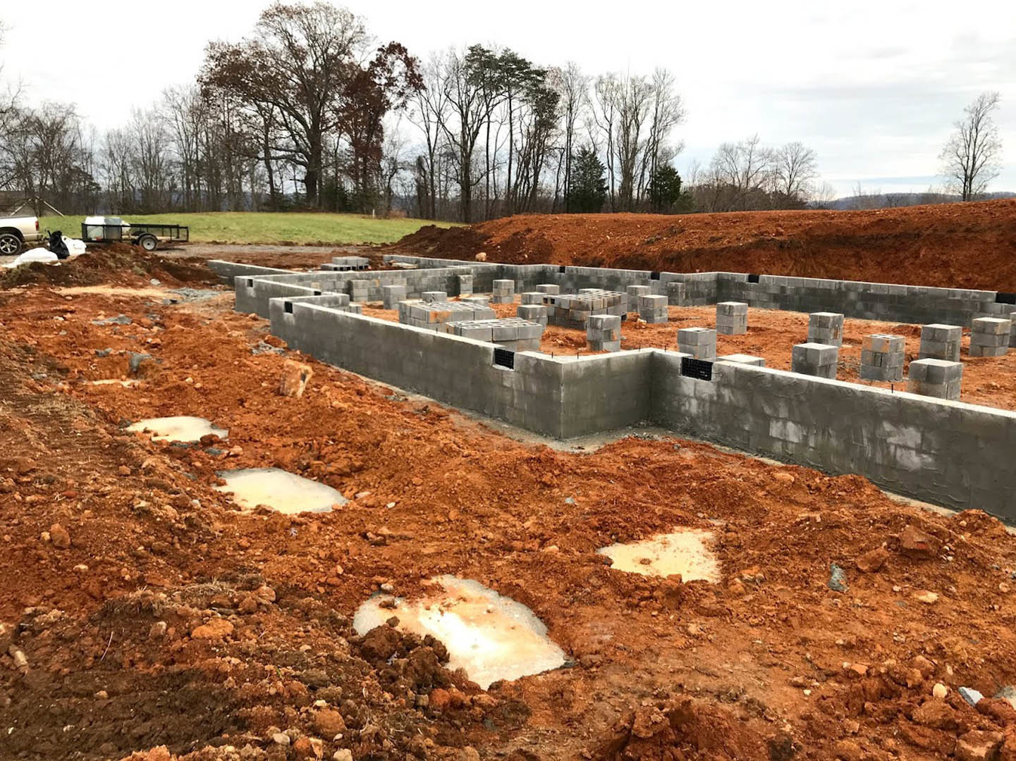 Concrete foundation with block walls set in excavated soil, surrounded by bare trees and patches of snow, white pickup truck parked nearby