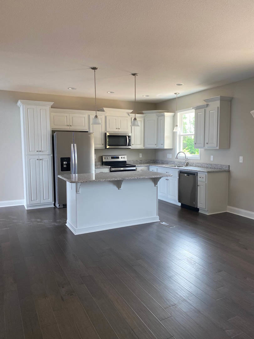 White kitchen with granite island, stainless steel refrigerator, built-in microwave, and modern stove, surrounded by light cabinetry and hardwood flooring.