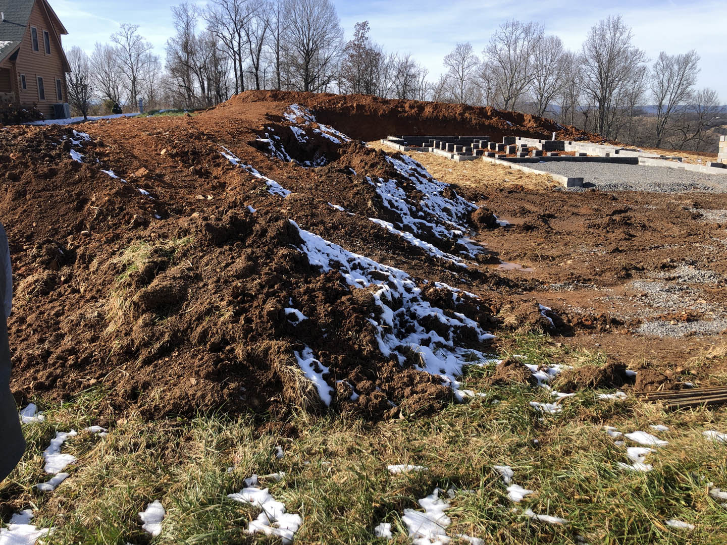 Modern home with dark roof and large windows set atop a hill, foreground features a mound of dirt and snow with patches of grass, winter landscape with bare trees and cloudy sky