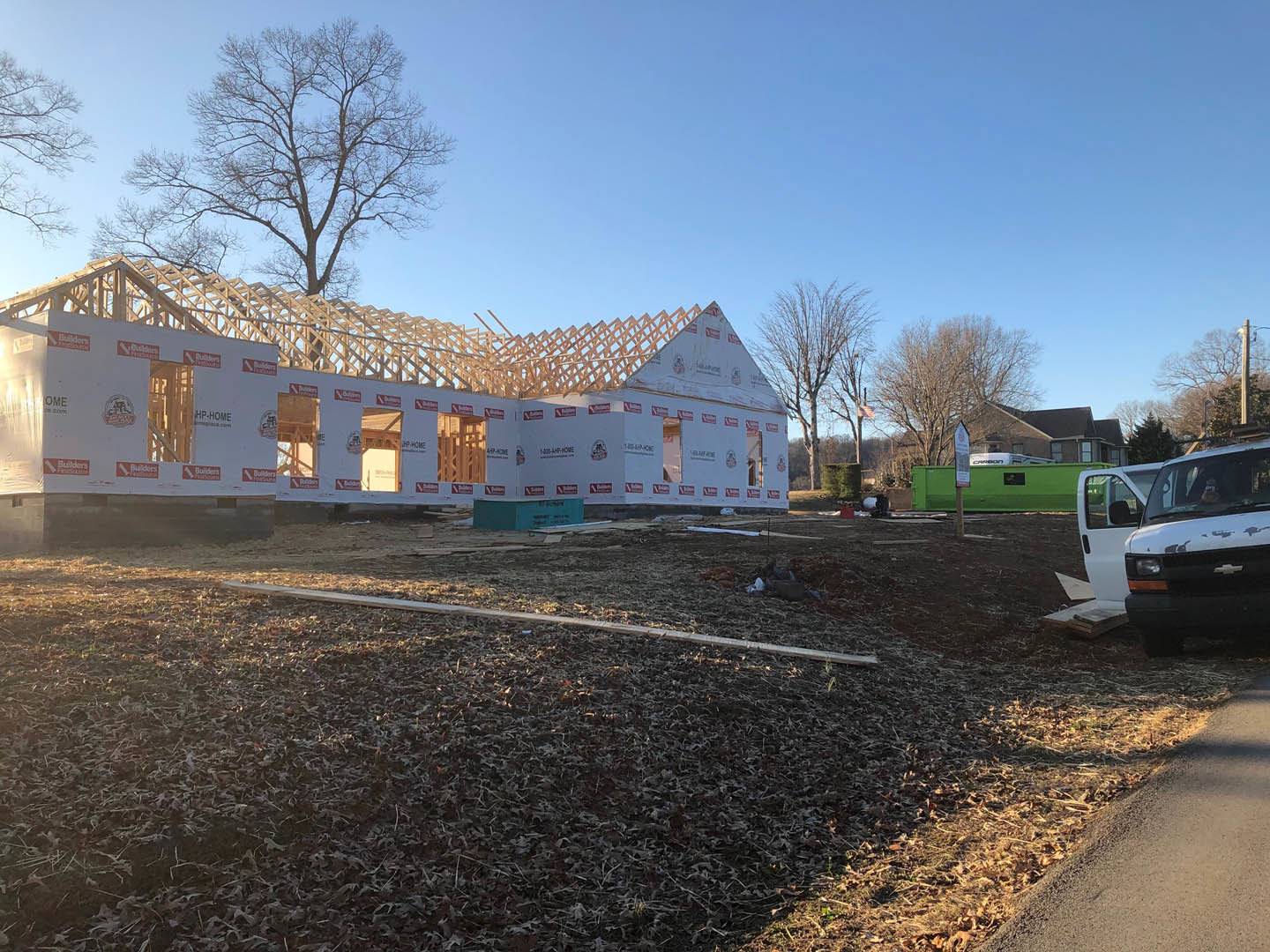 Partially built house with exposed framing and unfinished windows, green pickup truck parked in front, leafless tree and scattered leaves on the ground, wooden fence visible in