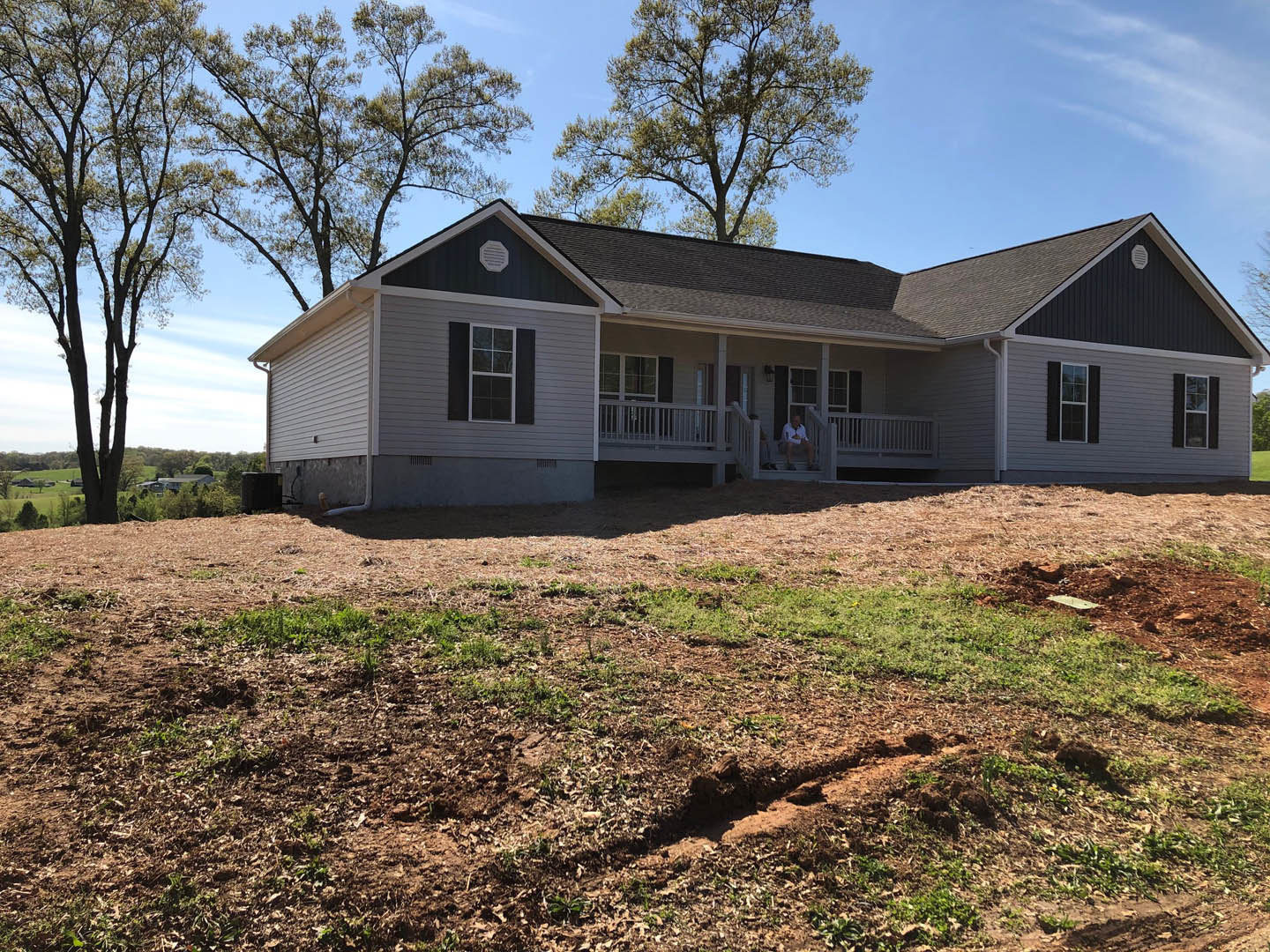 White-framed window and covered porch on a cottage-style house, two men seated on porch, large dirt patch with sparse grass in foreground, black container near entrance, trees in