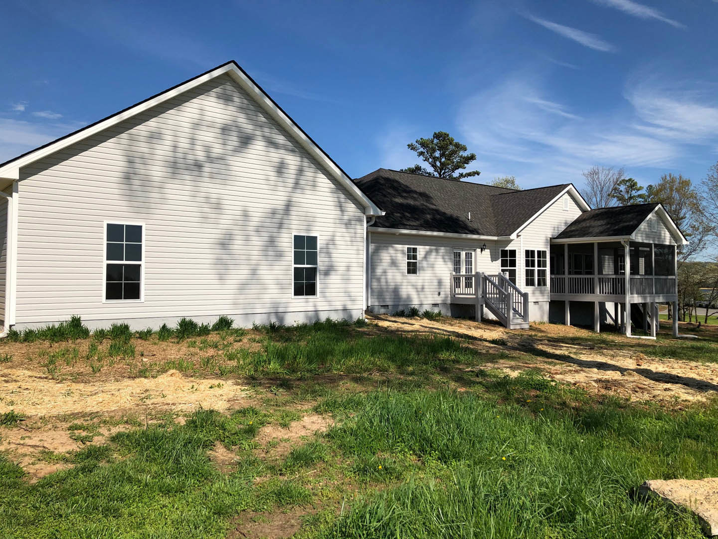 White house with multiple square windows, wooden porch with railings, green lawn in front, tree shadow cast on siding, cloudy sky overhead