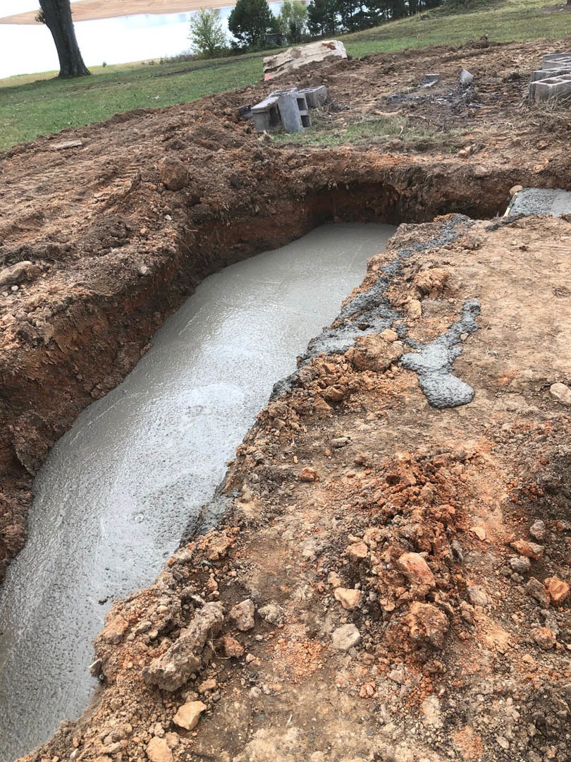 Cement slab foundation set in a dirt excavation, surrounded by grass, soil, and scattered concrete blocks, with trees and a water-filled ditch in the background